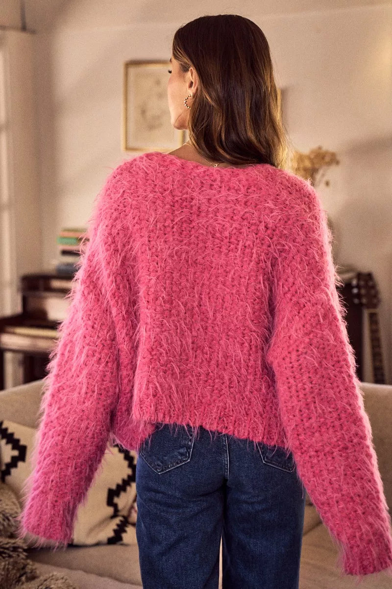 Woman wearing a fluffy pink oversized button-up cardigan layered over a white top in a cozy living room setting.