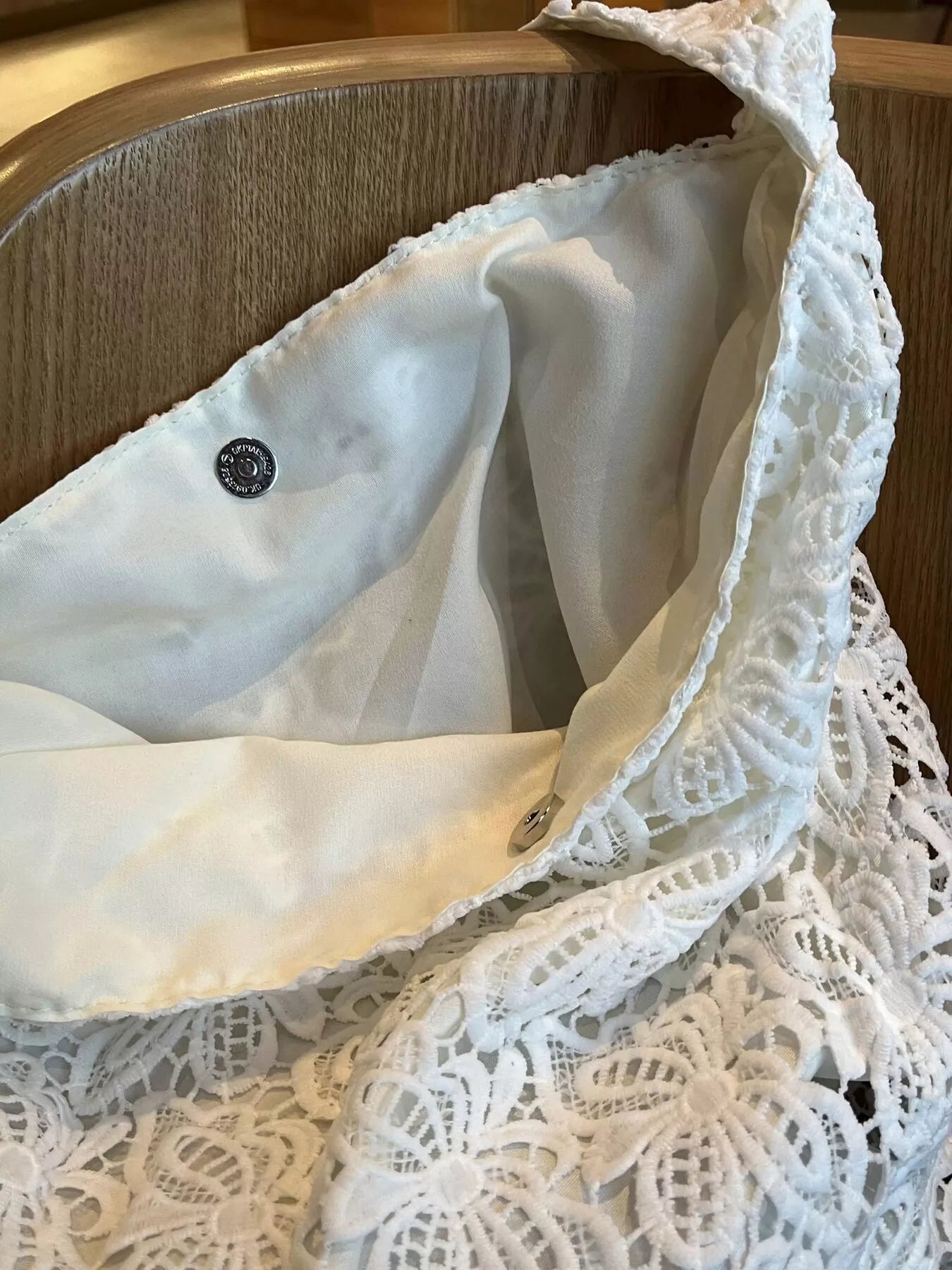 White medium-sized tote bag made of floral lace fabric with knotted straps, placed on a wooden chair in an indoor setting.