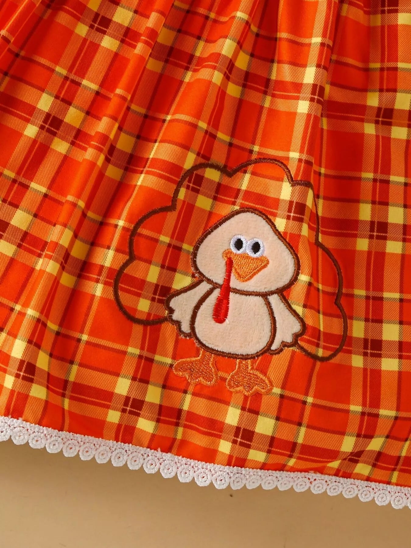 Three-piece baby girl outfit with orange plaid turkey dress, brown cardigan, and matching plaid headband on a beige background with fall leaves.