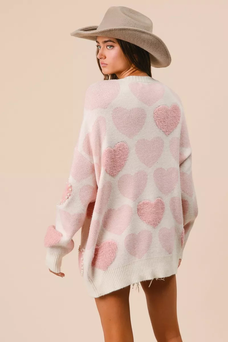 Woman wearing a cream knit sweater with pink heart pattern and pearl embellishments