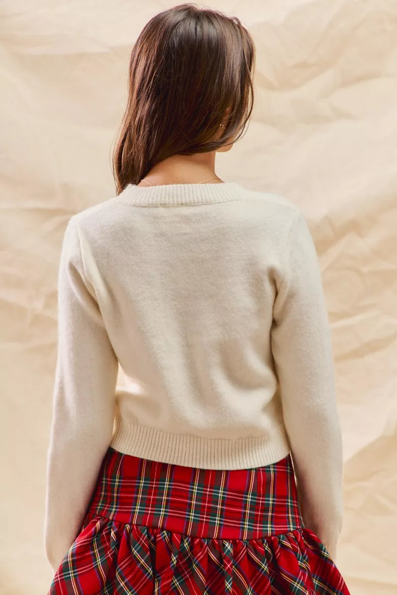 Woman wearing a cream cropped sweater with a red candy cane and holly design, paired with a red plaid skirt — festive holiday knit from A Whimsical Affair.