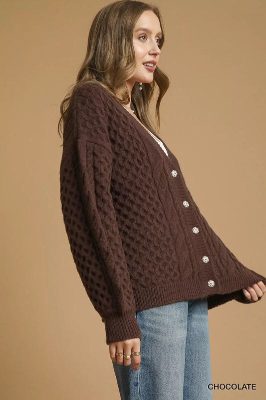 Model wearing a chocolate-brown cable-knit cardigan with pearl buttons, styled with a white tank top and jeans.