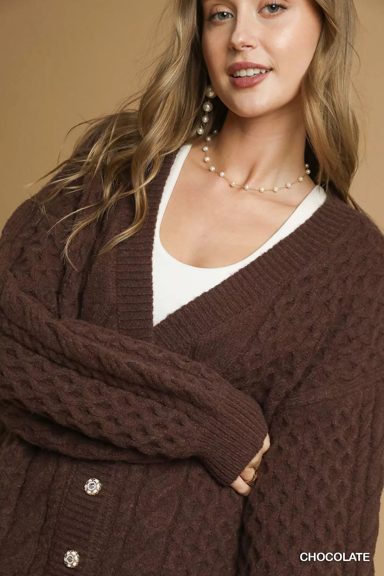 Model wearing a chocolate-brown cable-knit cardigan with pearl buttons, styled with a white tank top and jeans.