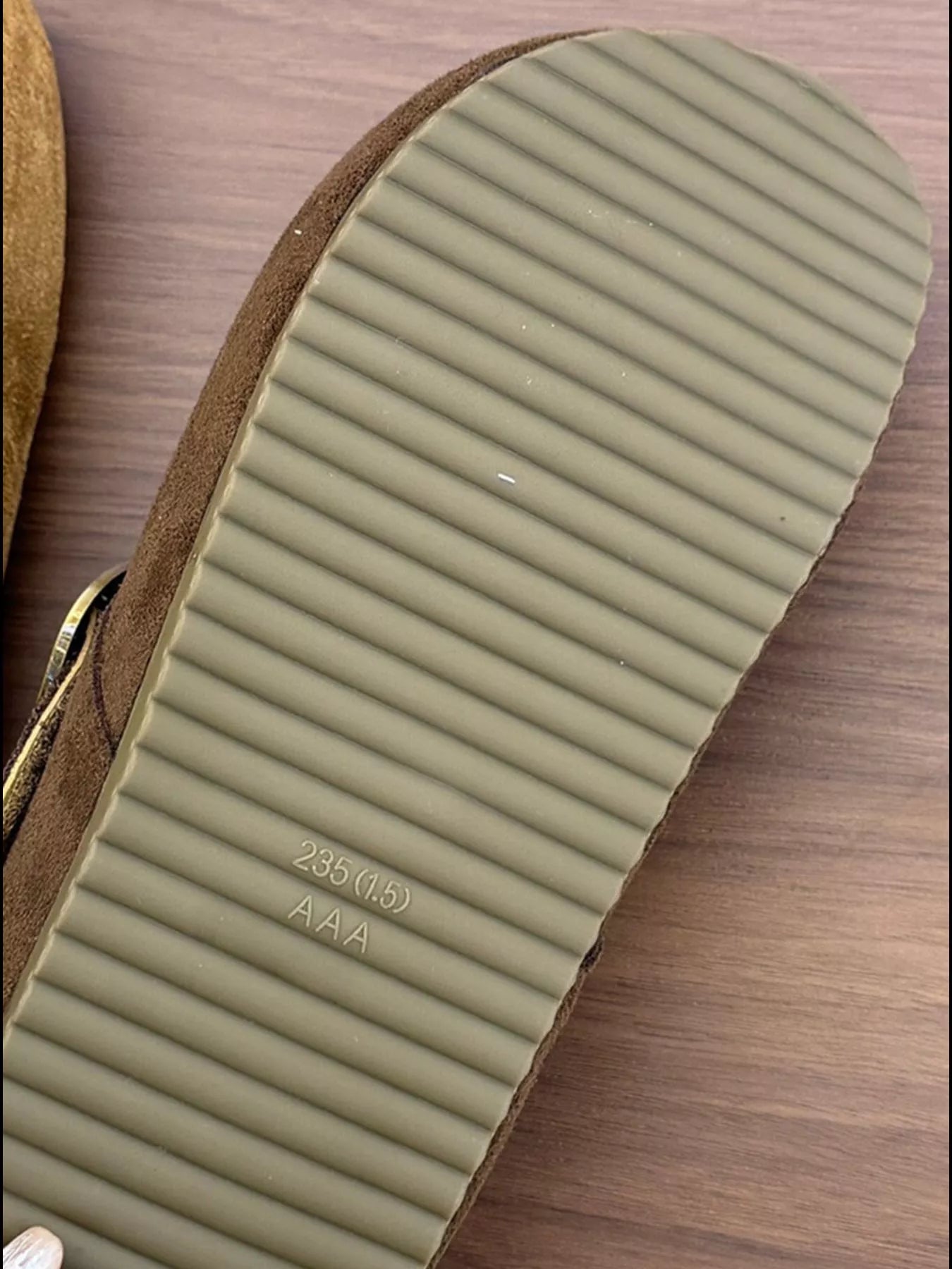 Pair of brown suede buckle clogs with a flat sole displayed on a wooden surface, showcasing the minimalist slip-on design.