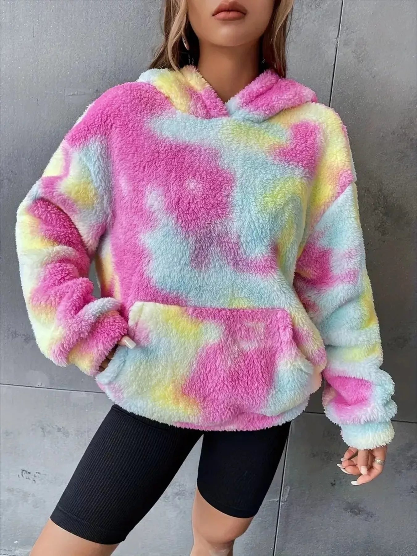 Model wearing a pastel tie-dye plush hoodie in pink, blue, and yellow with a kangaroo pocket and oversized cozy fit.