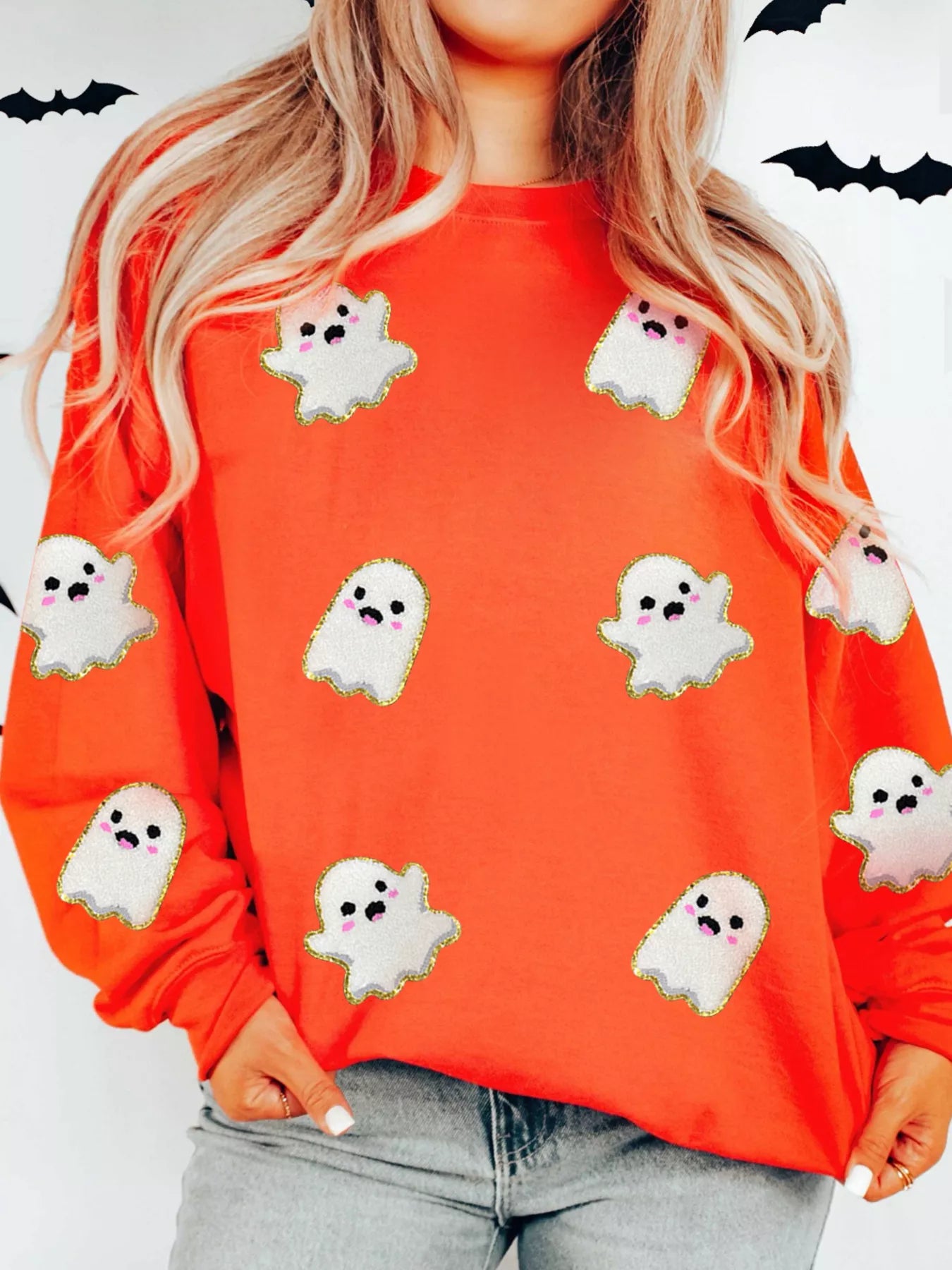 Woman wearing a bright orange sweatshirt decorated with cute white ghosts outlined in gold — festive Halloween sweatshirt from A Whimsical Affair.