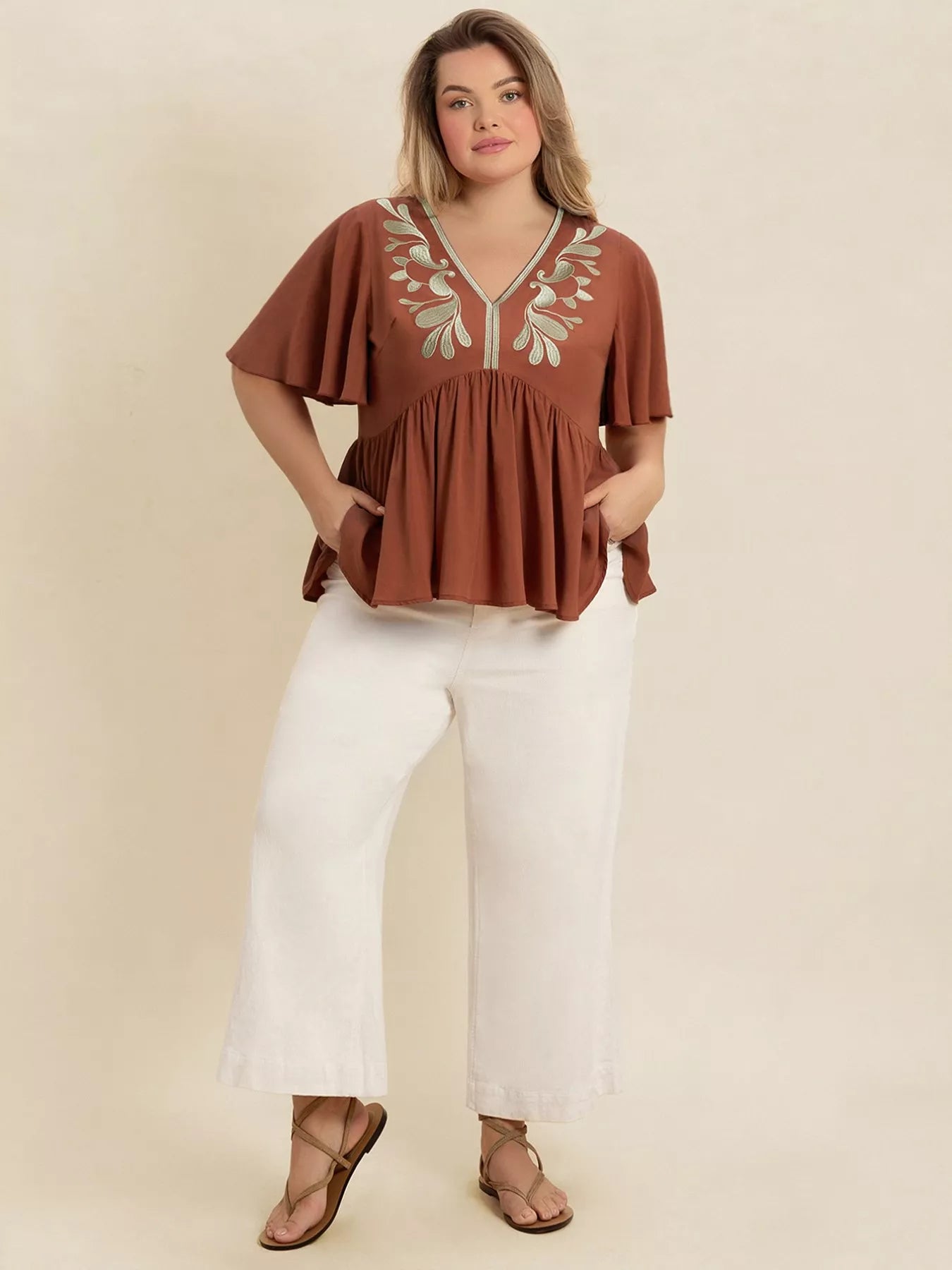 A woman wearing a rust-colored V-neck plus size blouse with embroidered floral detailing near the neckline, short flutter sleeves, and a ruched waist design.