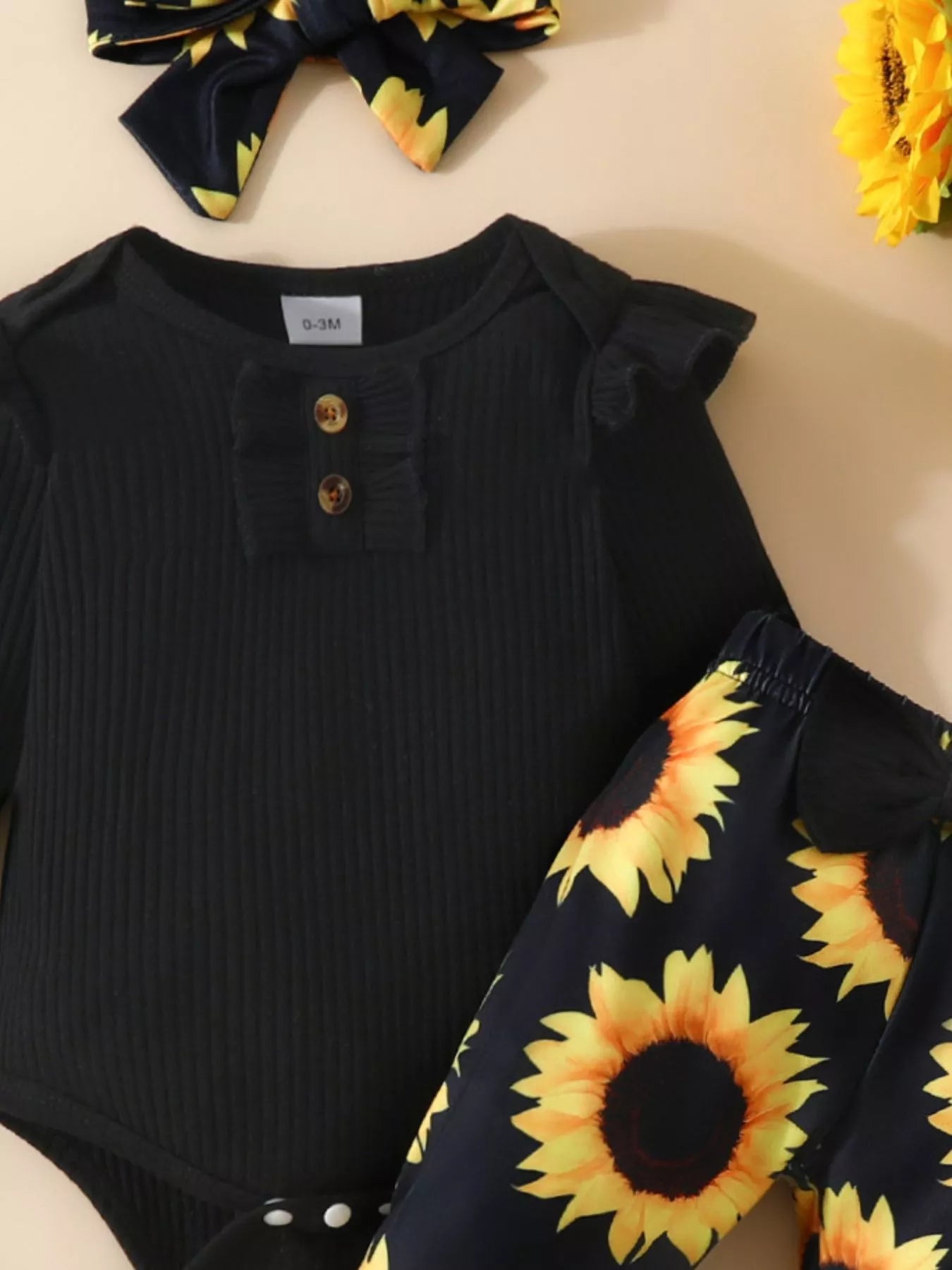 Baby outfit featuring a black ruffled romper, sunflower bell-bottom pants, and matching sunflower bow headband styled with floral decor.