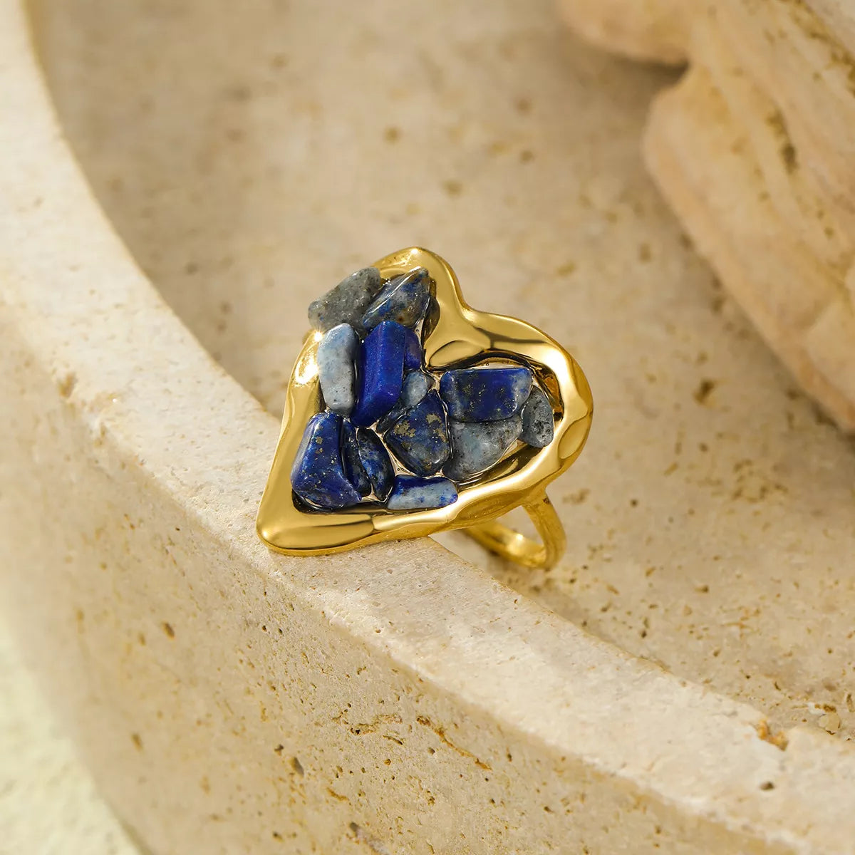 Gold heart ring with navy and slate lapis lazuli stones, styled against warm stone tones.