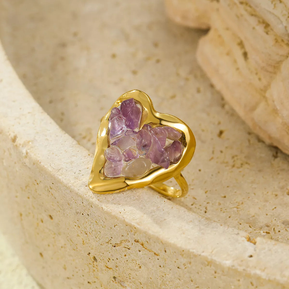 Gold heart ring filled with lilac amethyst stones on a neutral stone surface.