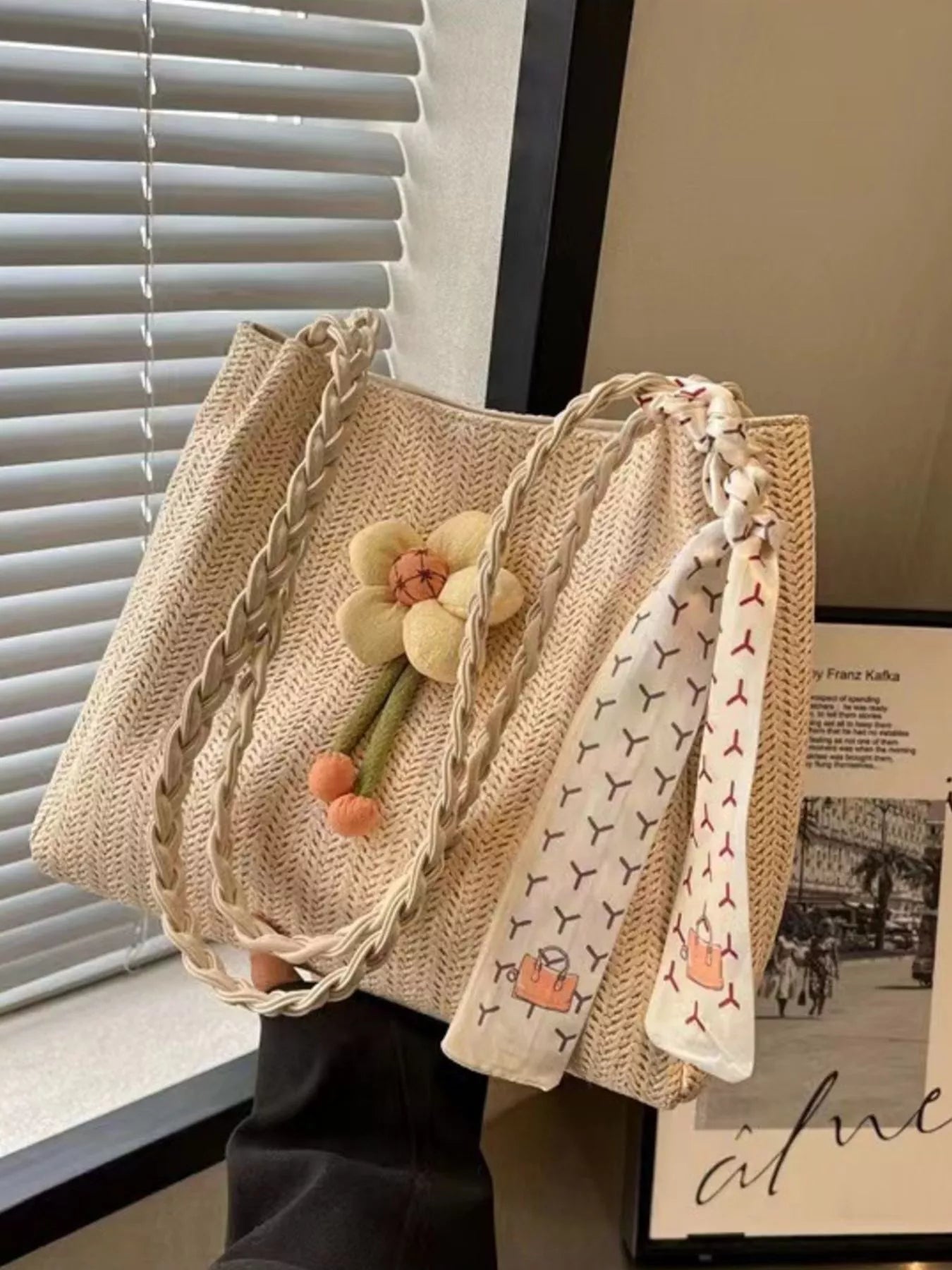 Beige woven straw tote bag with braided handles, a plush decorative flower, and a printed scarf detail