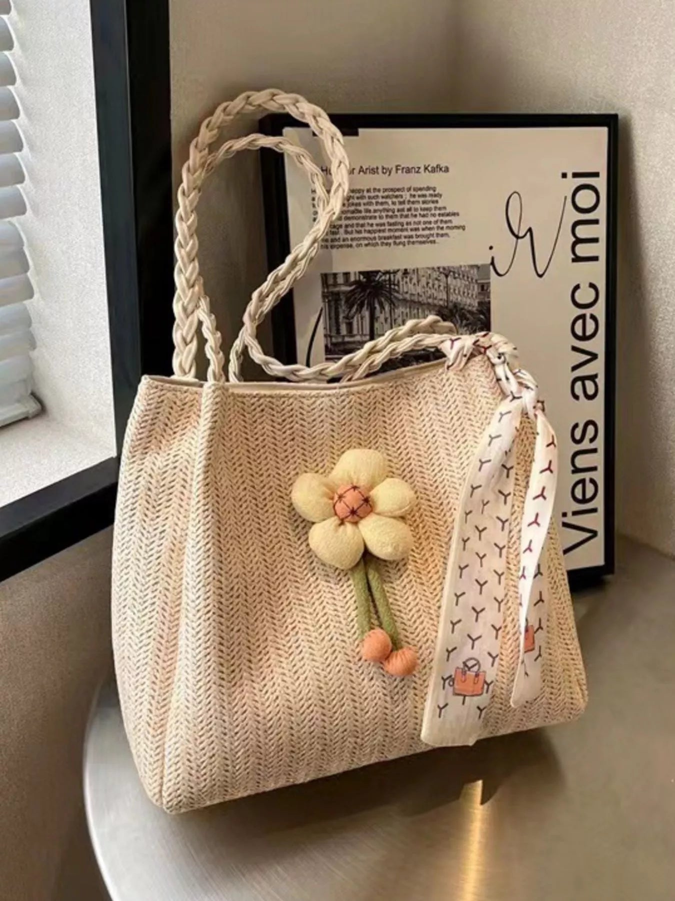 Beige woven straw tote bag with braided handles, a plush decorative flower, and a printed scarf detail