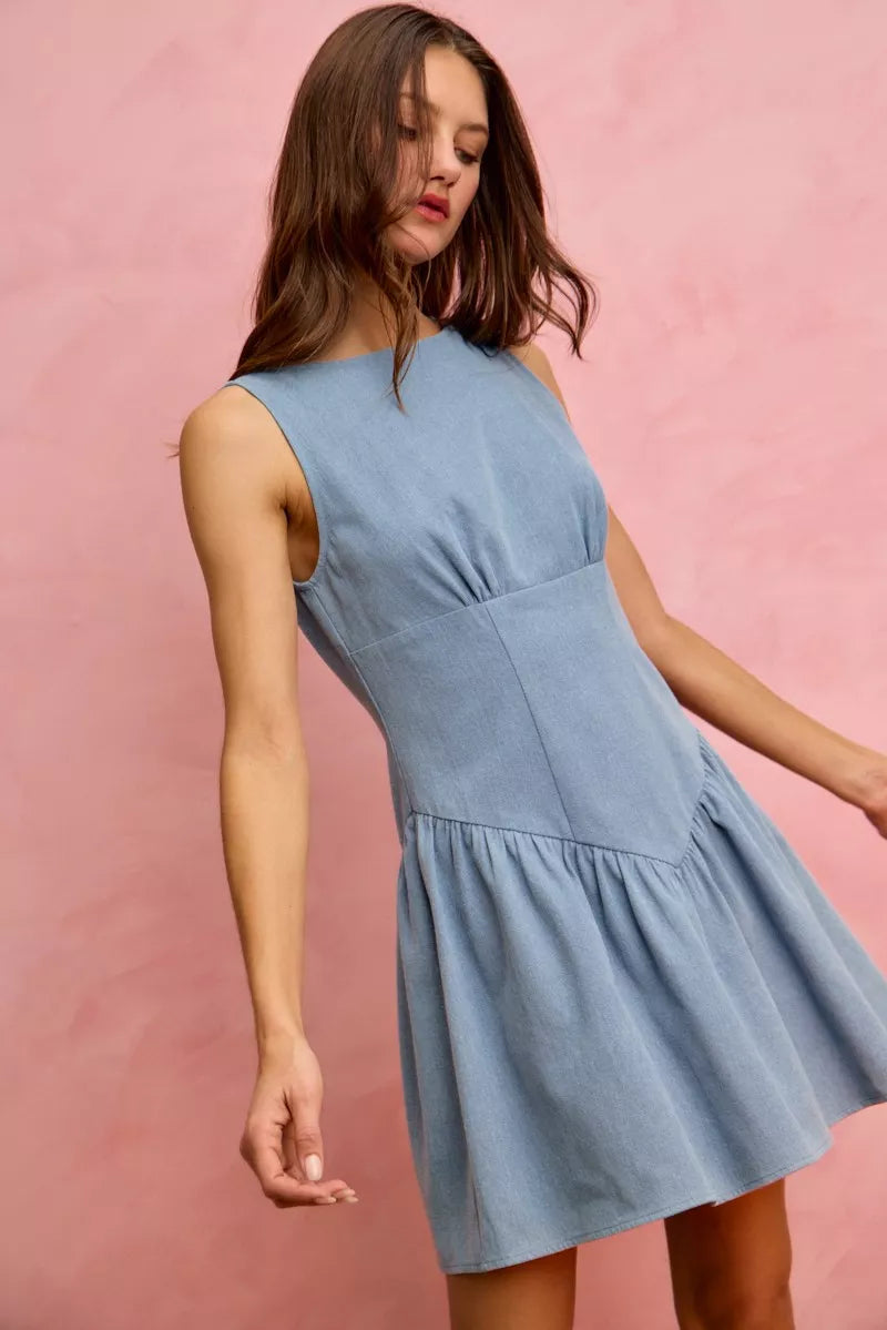 Model wearing a sleeveless fit and flare denim mini dress with structured bodice and flared skirt against a pink backdrop.