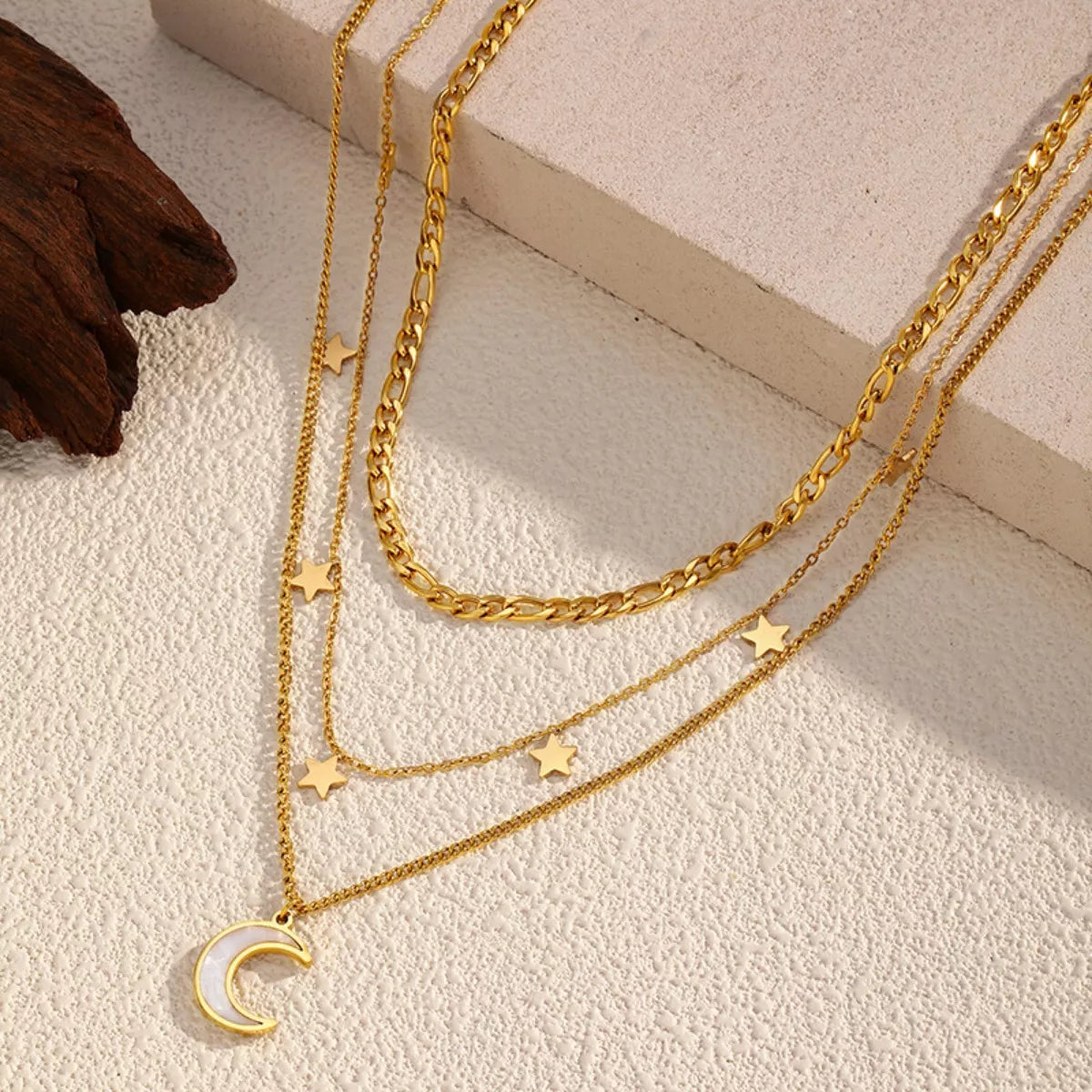 Gold layered necklace with three chains featuring small star charms and a crescent moon pendant displayed on a textured beige background.