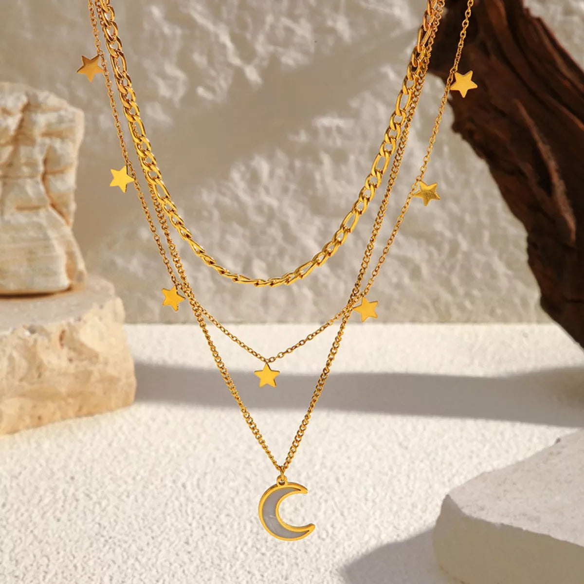 Gold layered necklace with three chains featuring small star charms and a crescent moon pendant displayed on a textured beige background.