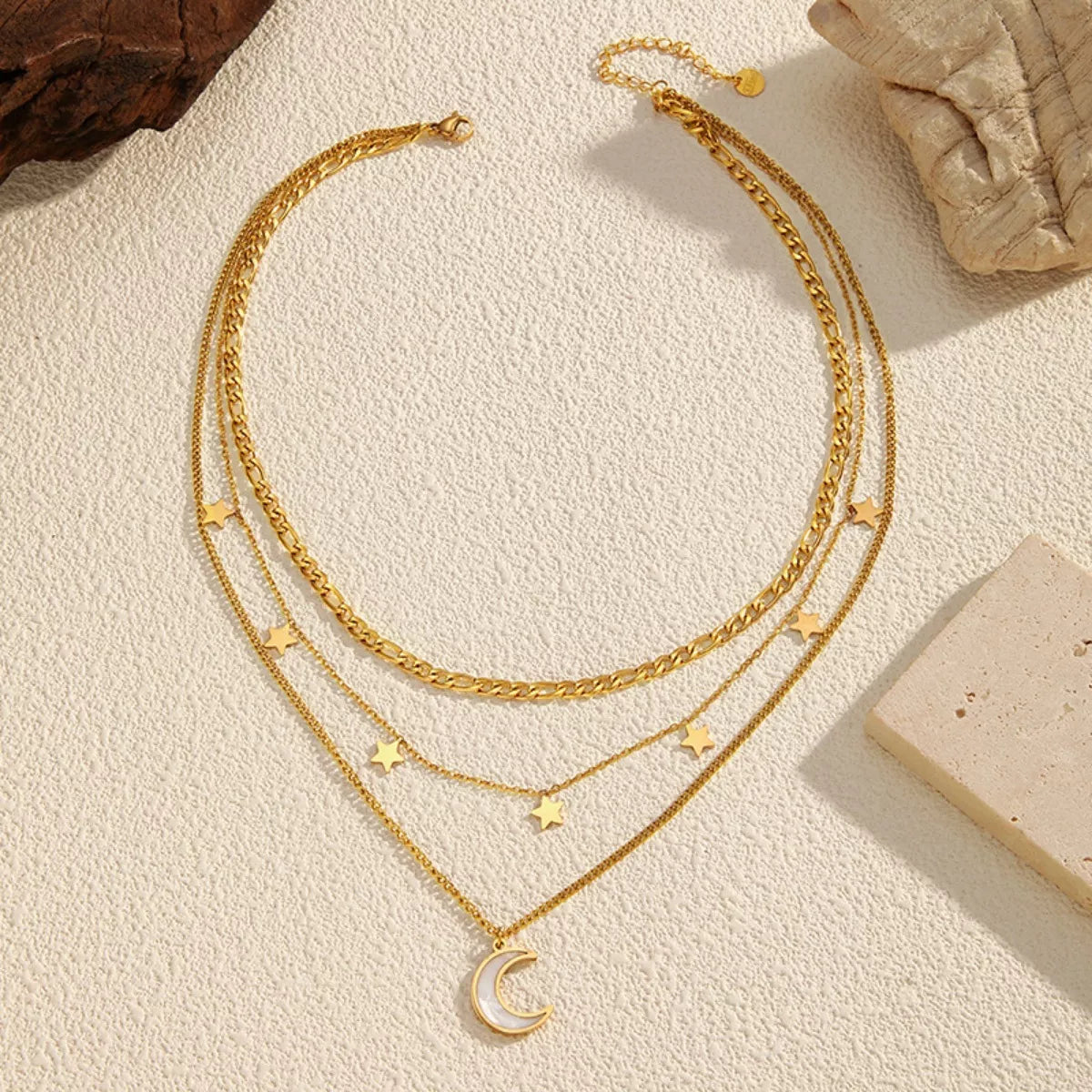 Gold layered necklace with three chains featuring small star charms and a crescent moon pendant displayed on a textured beige background.