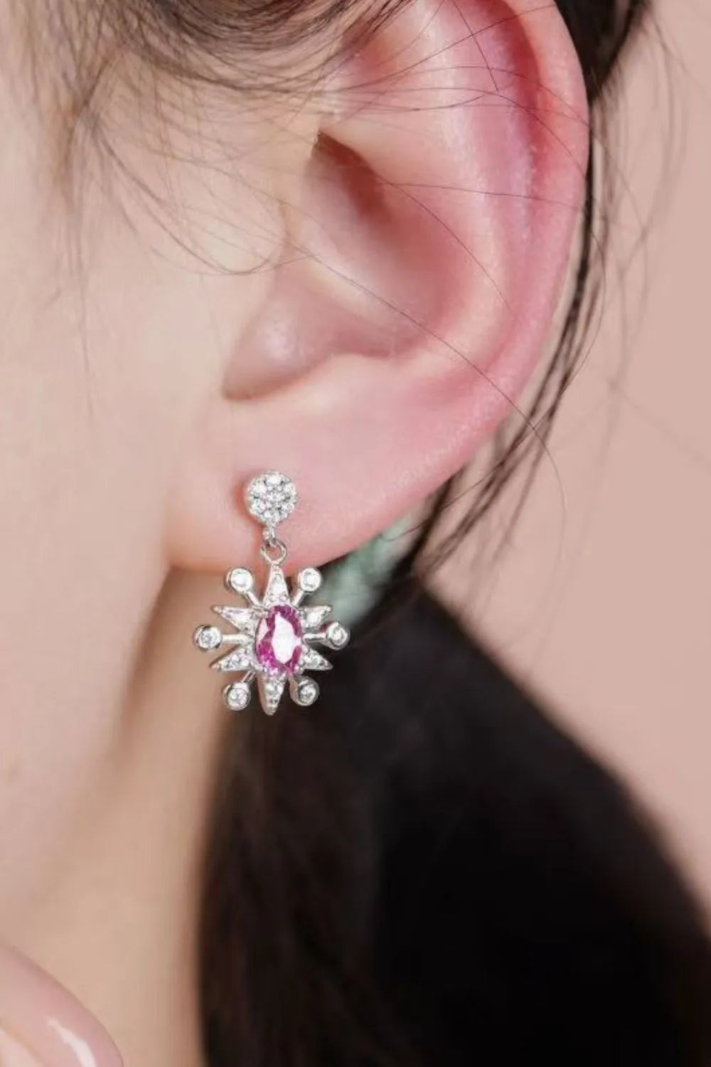 Close-up of mismatched sterling silver earrings featuring a crescent moon and a starburst, each with sparkling pink moissanite gemstones and crystal accents.