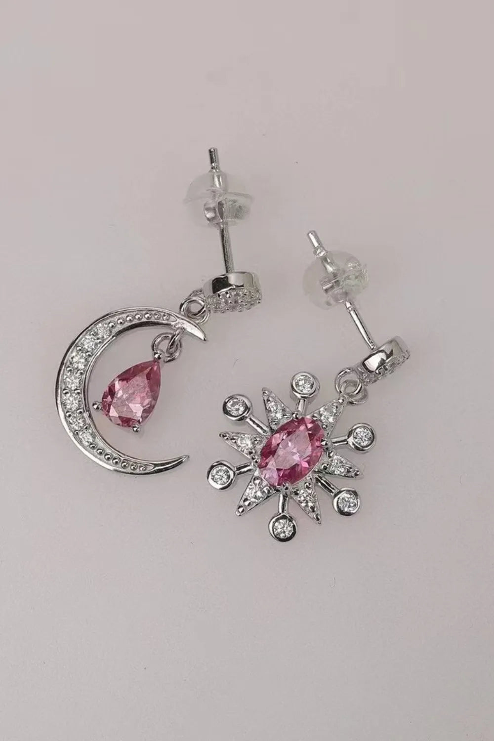 Close-up of mismatched sterling silver earrings featuring a crescent moon and a starburst, each with sparkling pink moissanite gemstones and crystal accents.