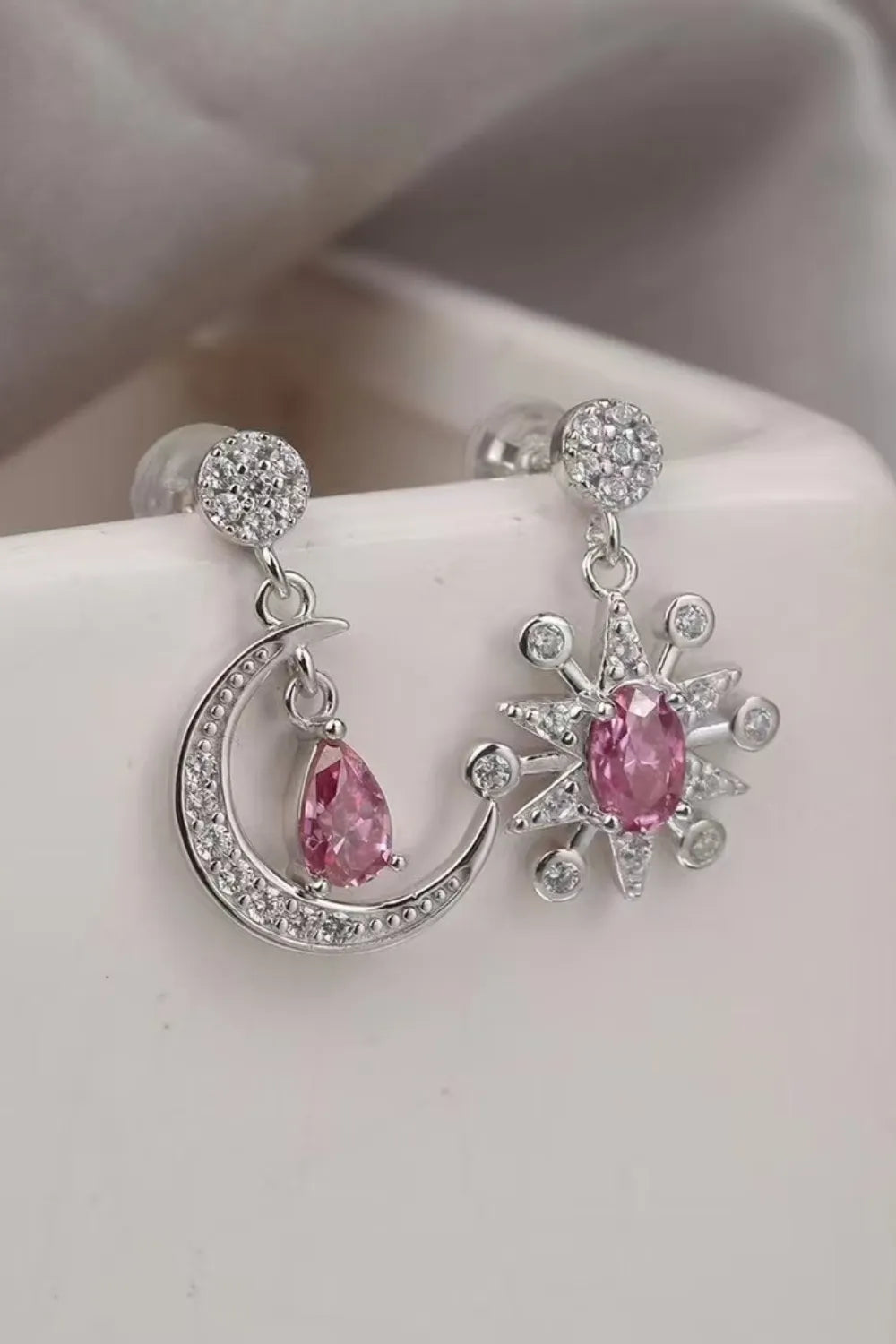 Close-up of mismatched sterling silver earrings featuring a crescent moon and a starburst, each with sparkling pink moissanite gemstones and crystal accents.