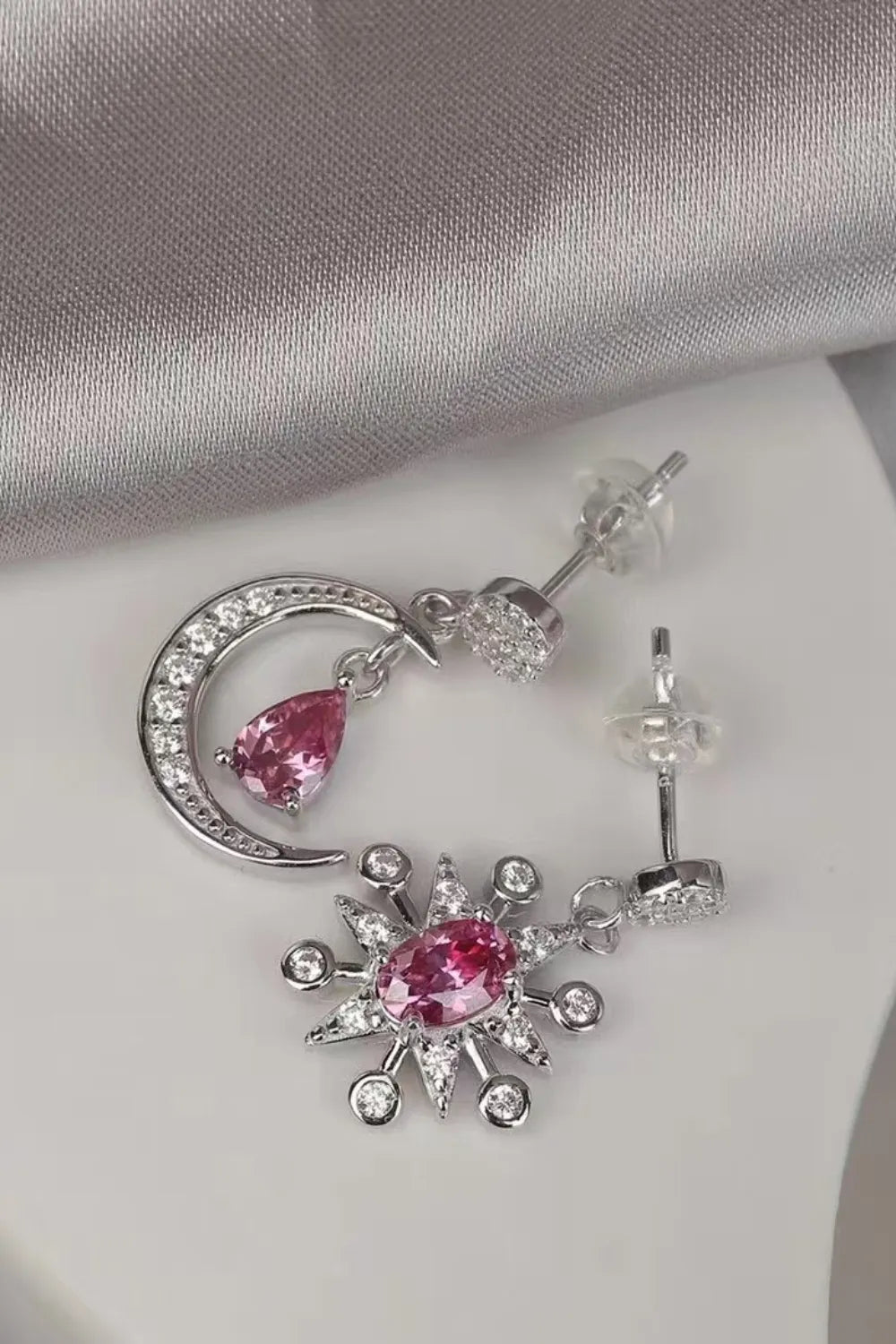 Close-up of mismatched sterling silver earrings featuring a crescent moon and a starburst, each with sparkling pink moissanite gemstones and crystal accents.