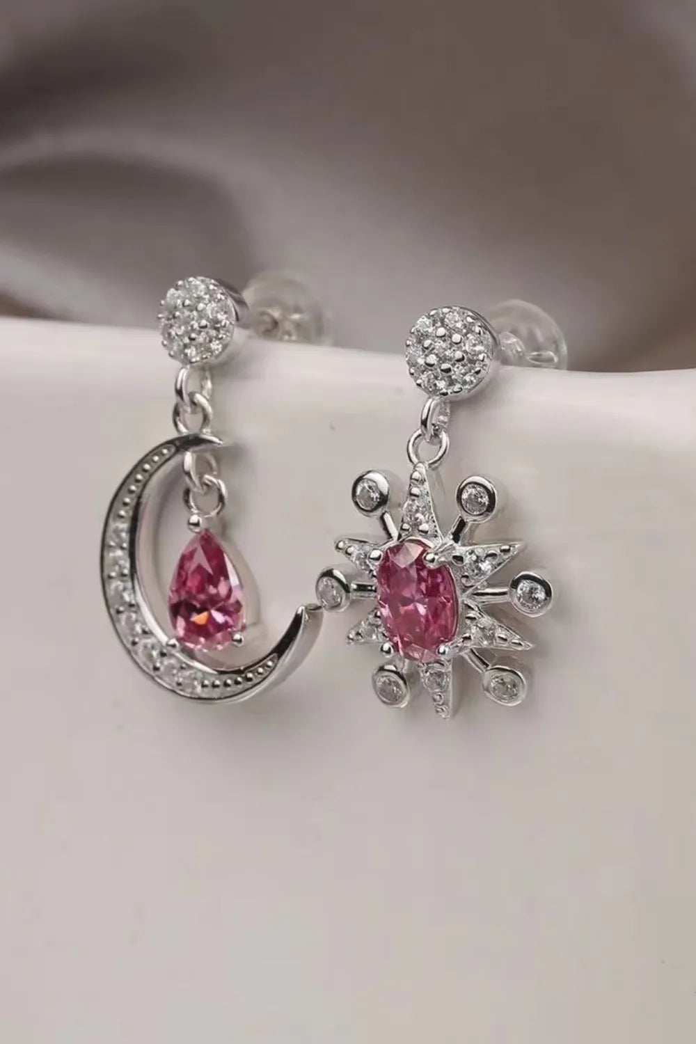 Close-up of mismatched sterling silver earrings featuring a crescent moon and a starburst, each with sparkling pink moissanite gemstones and crystal accents.