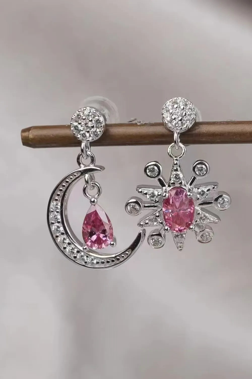 Close-up of mismatched sterling silver earrings featuring a crescent moon and a starburst, each with sparkling pink moissanite gemstones and crystal accents.