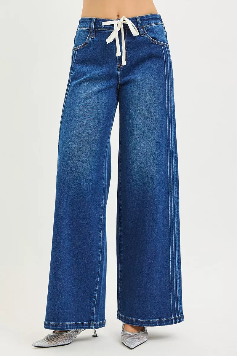 Woman wearing mid-rise wide leg blue jeans with drawstring waist and pintuck seams, standing against a neutral background.