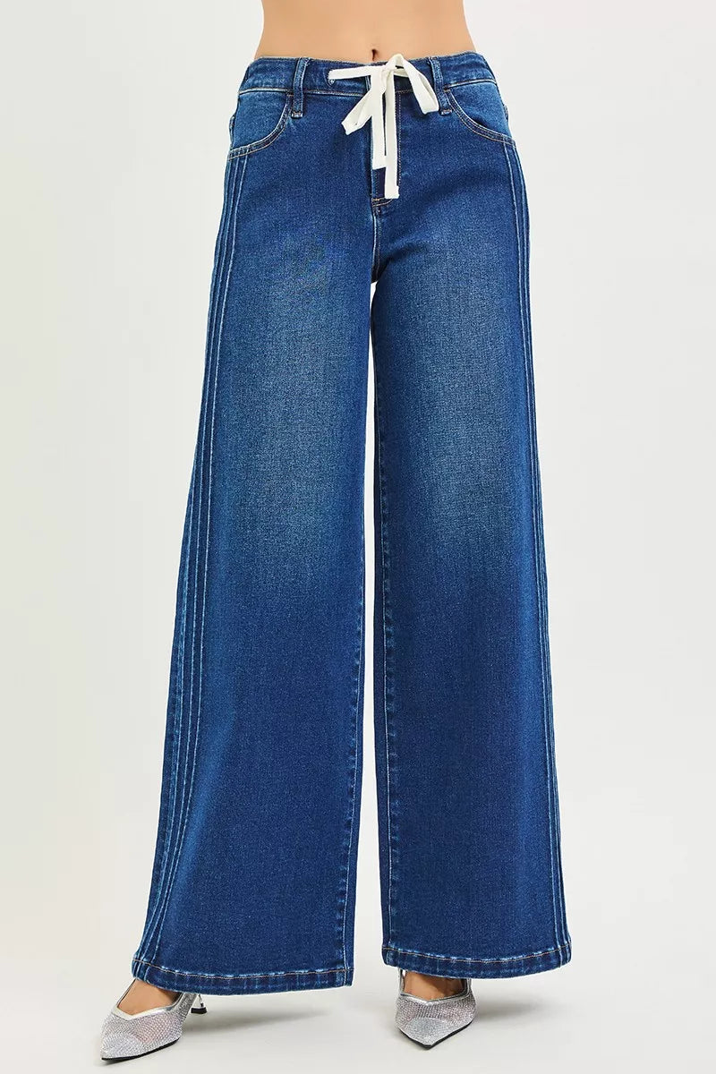 Woman wearing mid-rise wide leg blue jeans with drawstring waist and pintuck seams, standing against a neutral background.