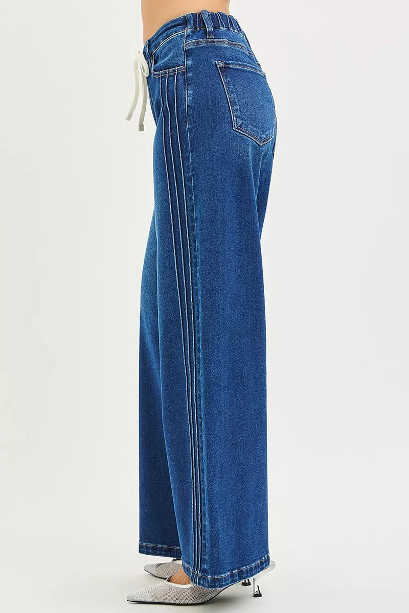 Woman wearing mid-rise wide leg blue jeans with drawstring waist and pintuck seams, standing against a neutral background.