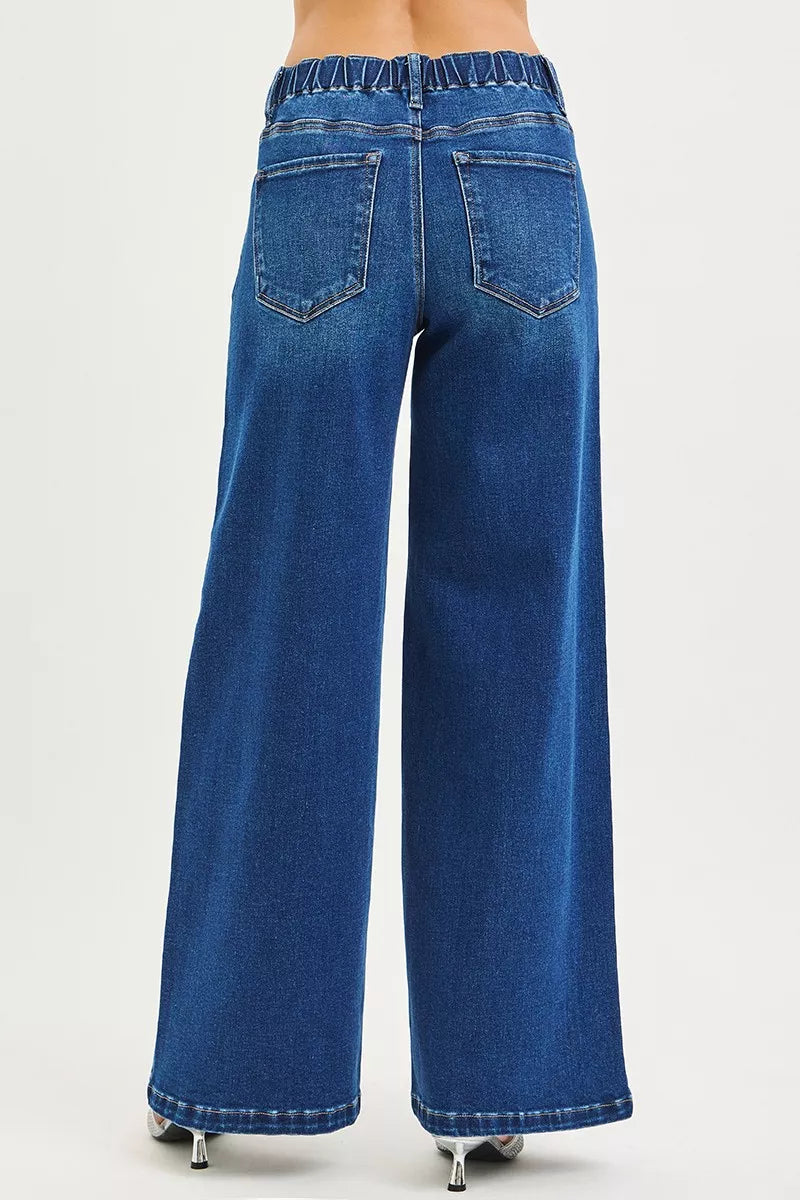 Woman wearing mid-rise wide leg blue jeans with drawstring waist and pintuck seams, standing against a neutral background.