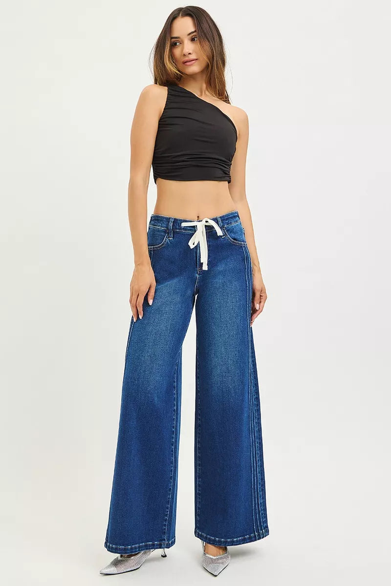 Woman wearing mid-rise wide leg blue jeans with drawstring waist and pintuck seams, standing against a neutral background.