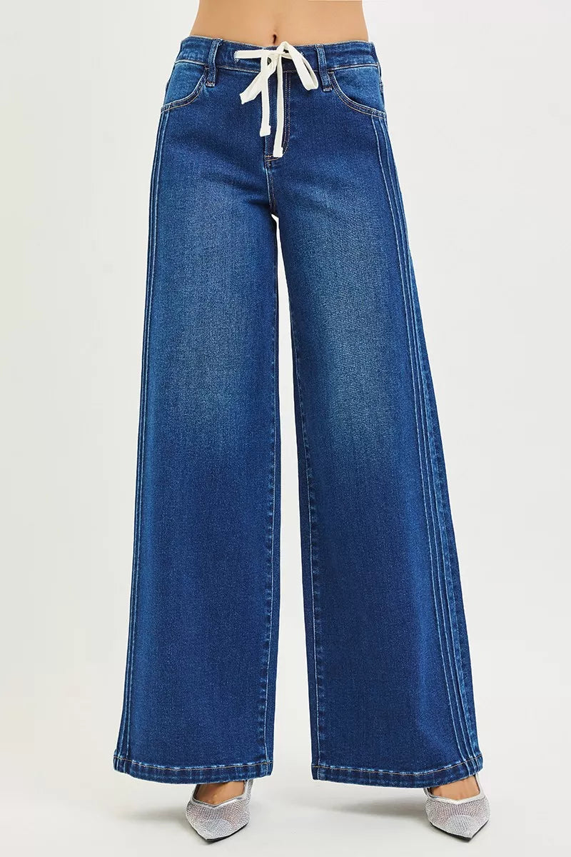 Woman wearing mid-rise wide leg blue jeans with drawstring waist and pintuck seams, standing against a neutral background.