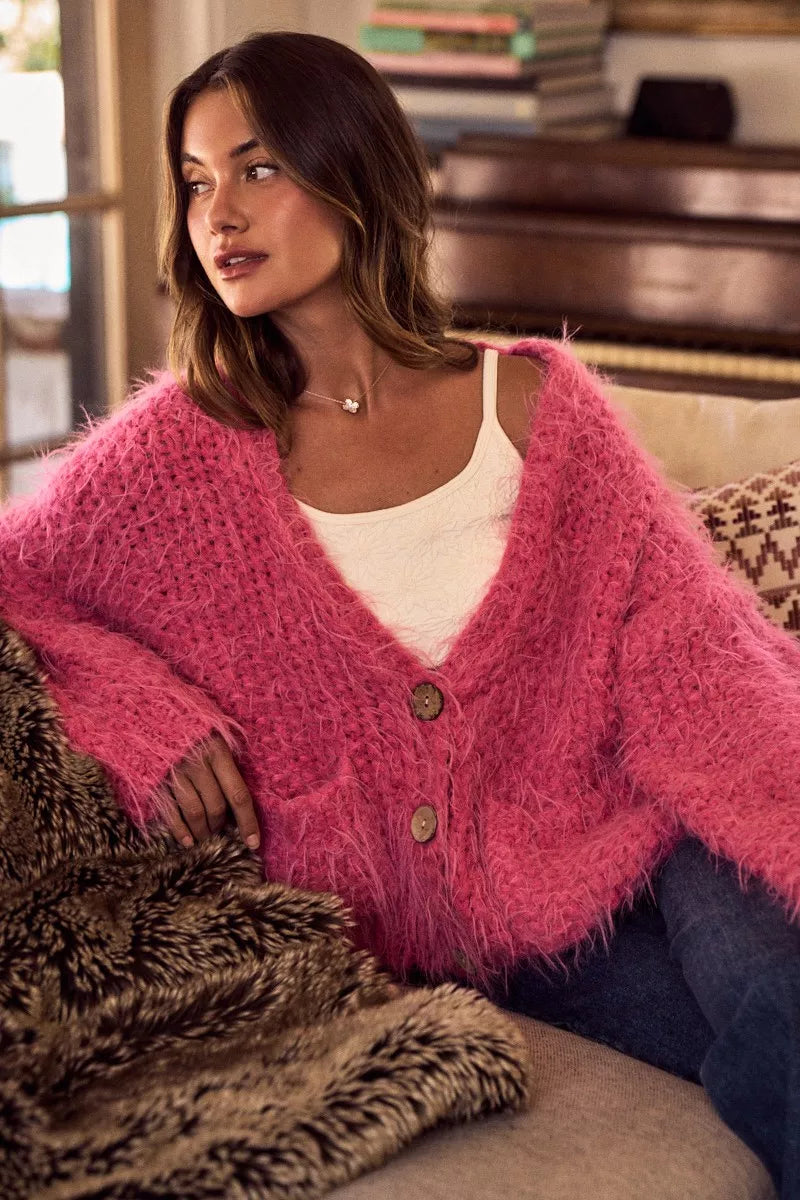 Woman wearing a fluffy pink oversized button-up cardigan layered over a white top in a cozy living room setting.