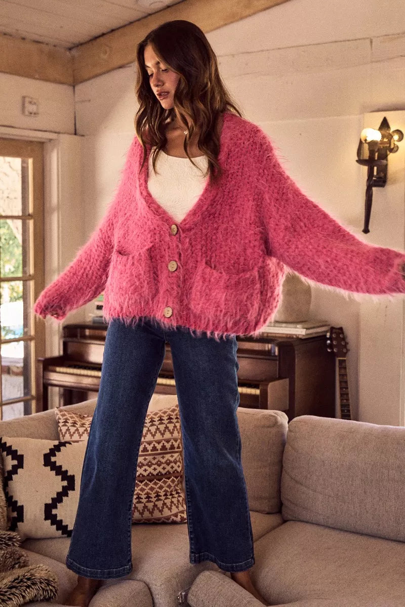 Woman wearing a fluffy pink oversized button-up cardigan layered over a white top in a cozy living room setting.