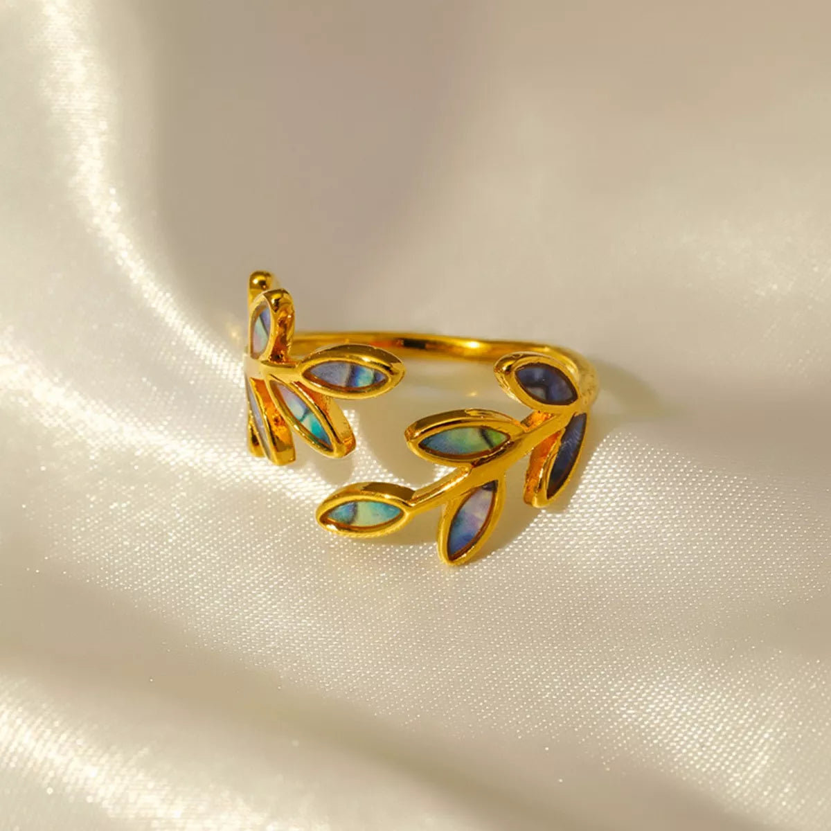 Gold-plated open ring featuring iridescent abalone leaf-shaped inlays displayed on soft cream satin fabric.