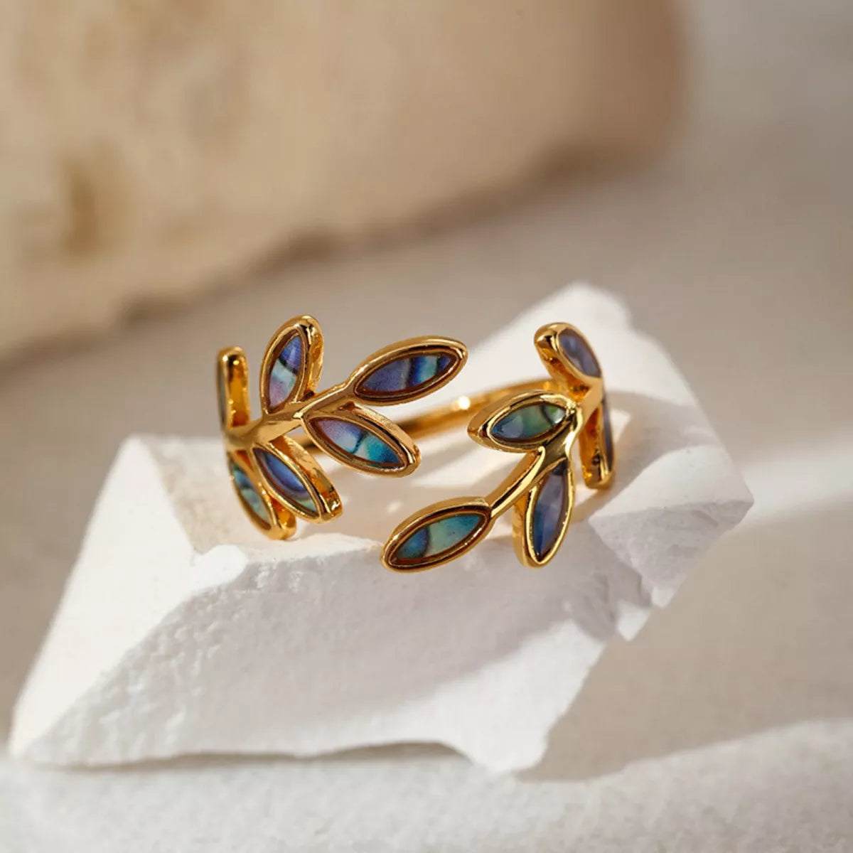 Gold-plated open ring featuring iridescent abalone leaf-shaped inlays displayed on soft cream satin fabric.