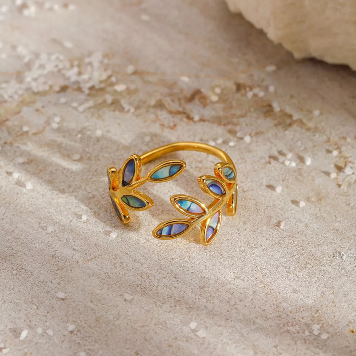 Gold-plated open ring featuring iridescent abalone leaf-shaped inlays displayed on soft cream satin fabric.