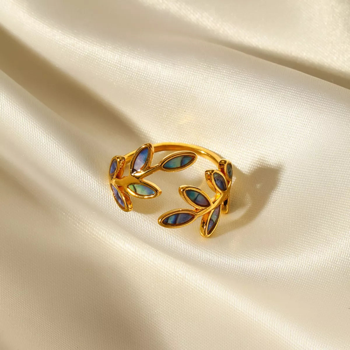 Gold-plated open ring featuring iridescent abalone leaf-shaped inlays displayed on soft cream satin fabric.