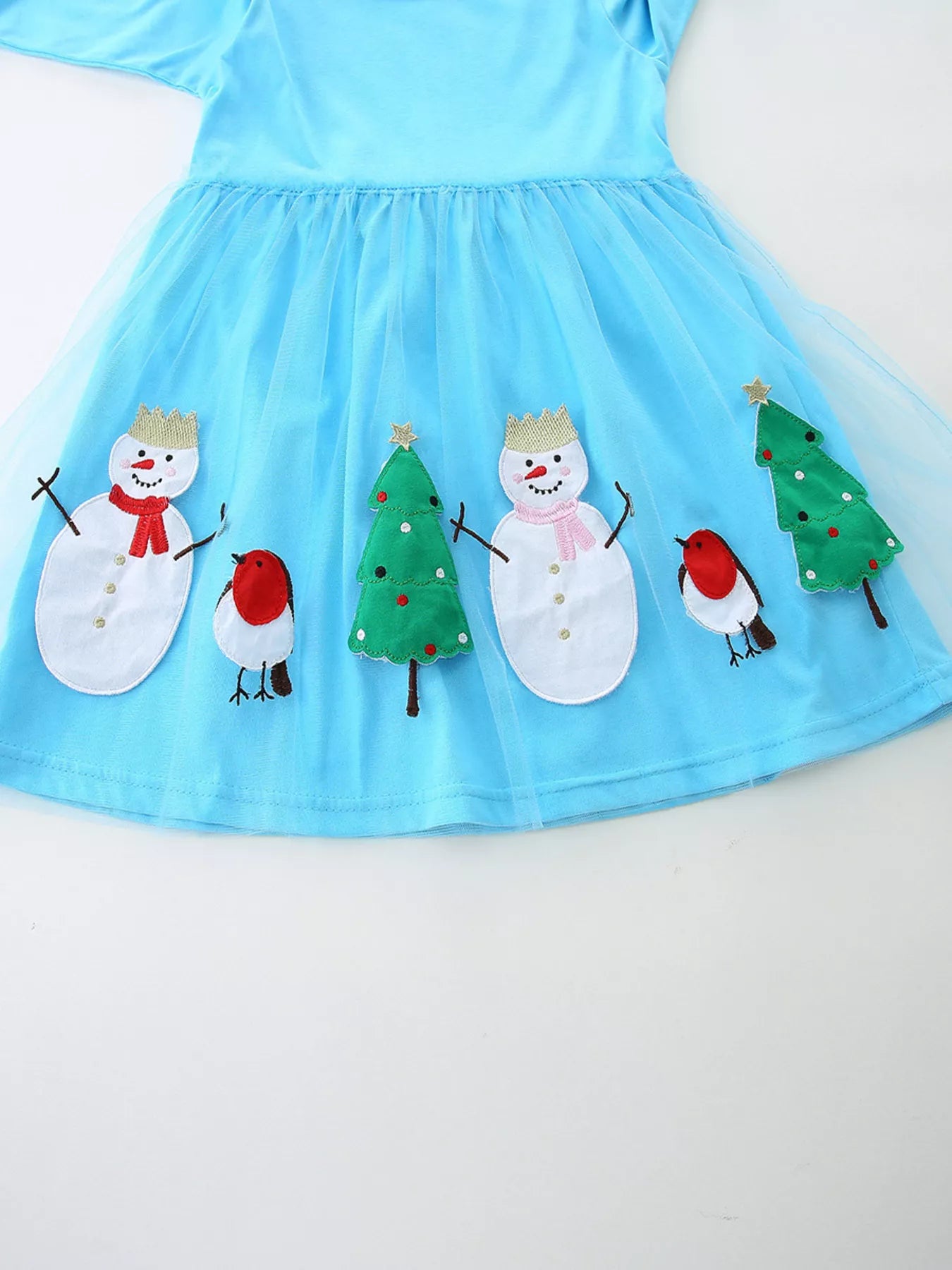A little girl wearing a light blue long-sleeve dress with snowman, Christmas tree, and bird appliqués on a mesh skirt overlay.