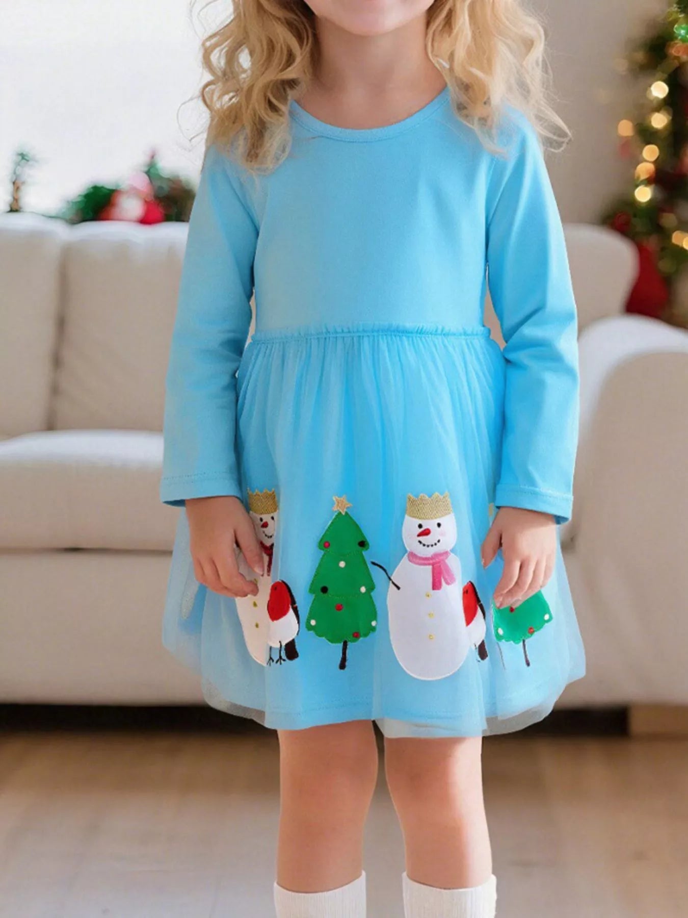 A little girl wearing a light blue long-sleeve dress with snowman, Christmas tree, and bird appliqués on a mesh skirt overlay.
