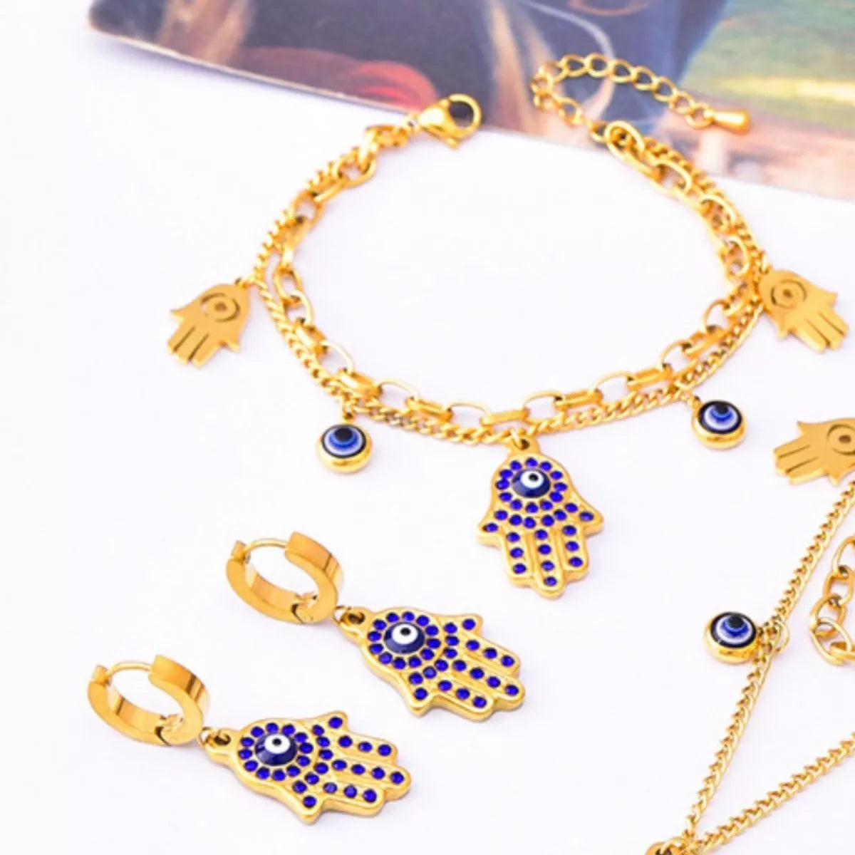 Gold-plated 3-piece jewelry set featuring a Hamsa hand charm bracelet, matching Hamsa earrings, and Evil Eye accents with blue zircon detailing on a white background.