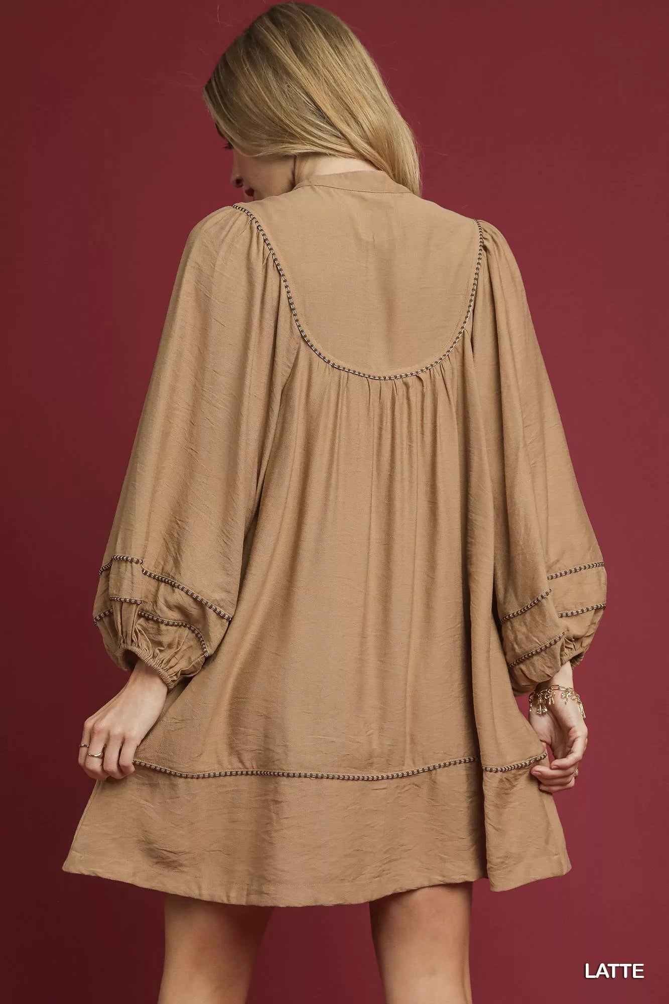 Woman wearing a latte-colored boho peasant mini dress with balloon sleeves, tassel ties, and stitched trim details against a burgundy background.