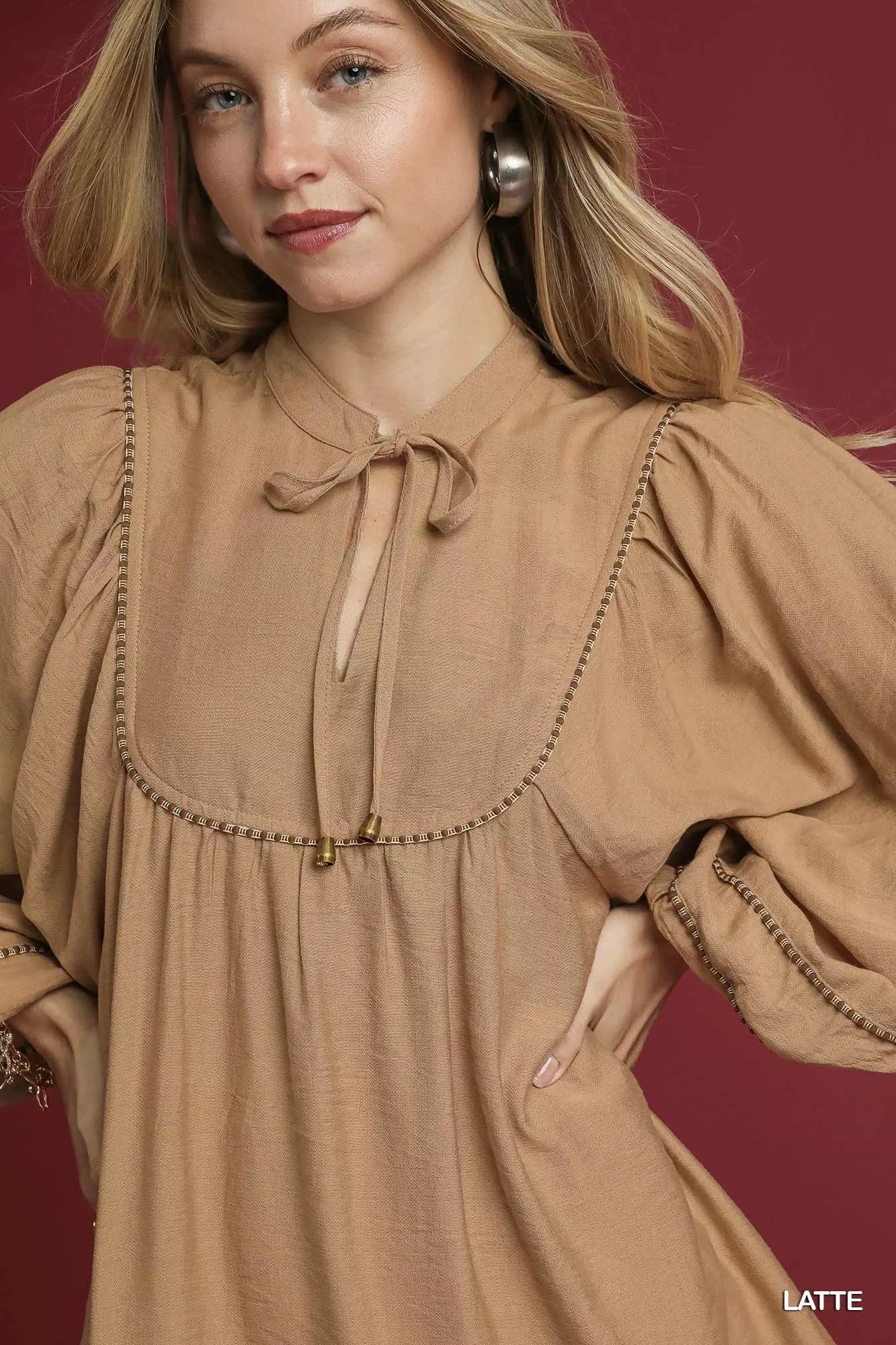 Woman wearing a latte-colored boho peasant mini dress with balloon sleeves, tassel ties, and stitched trim details against a burgundy background.