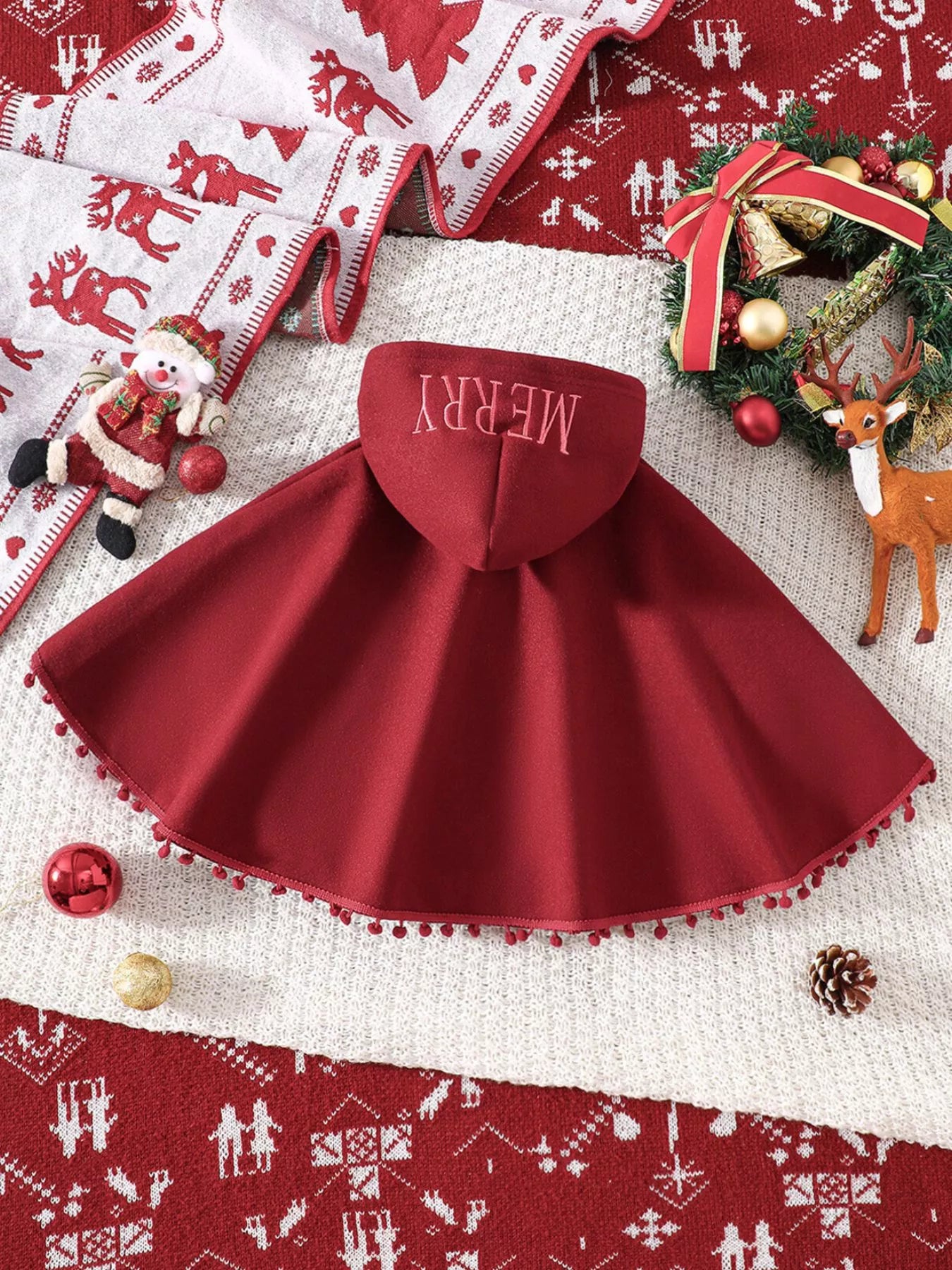 Red Christmas hooded cape for kids with “MERRY” embroidery and reindeer appliqués, pom-trim hem, displayed on holiday background.
