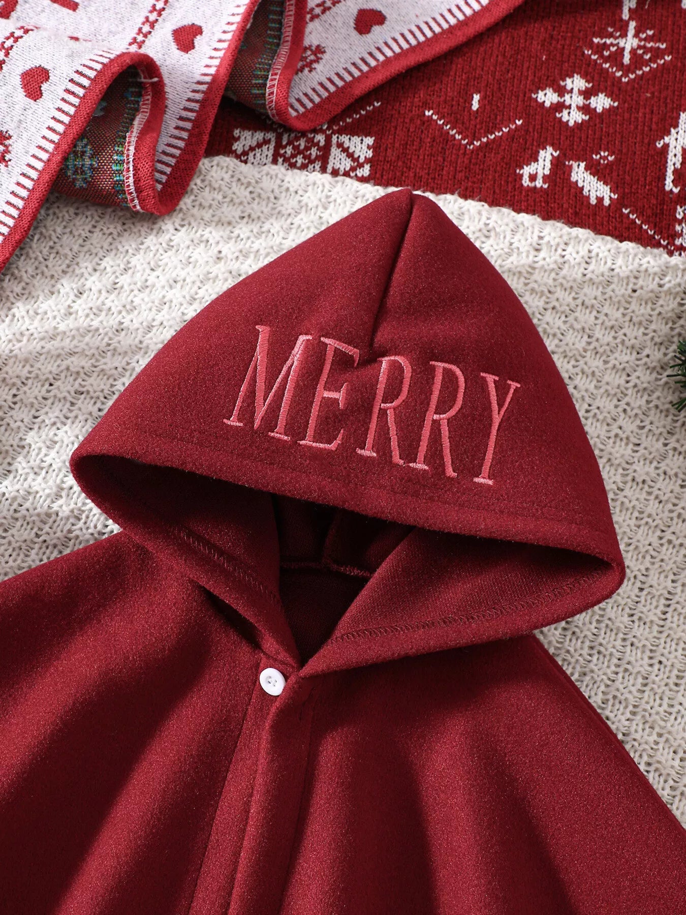 Red Christmas hooded cape for kids with “MERRY” embroidery and reindeer appliqués, pom-trim hem, displayed on holiday background.
