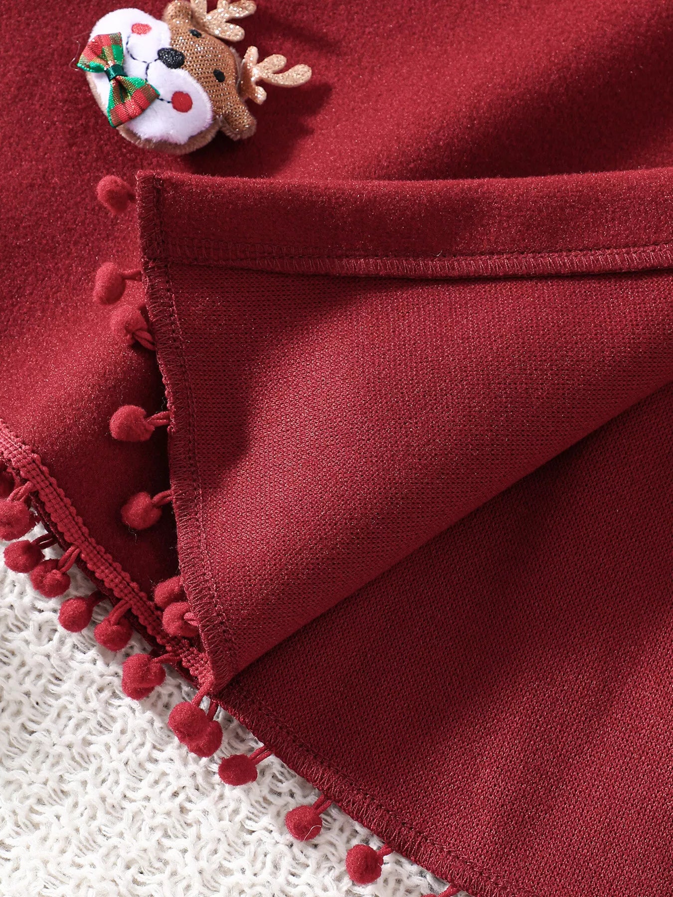 Red Christmas hooded cape for kids with “MERRY” embroidery and reindeer appliqués, pom-trim hem, displayed on holiday background.