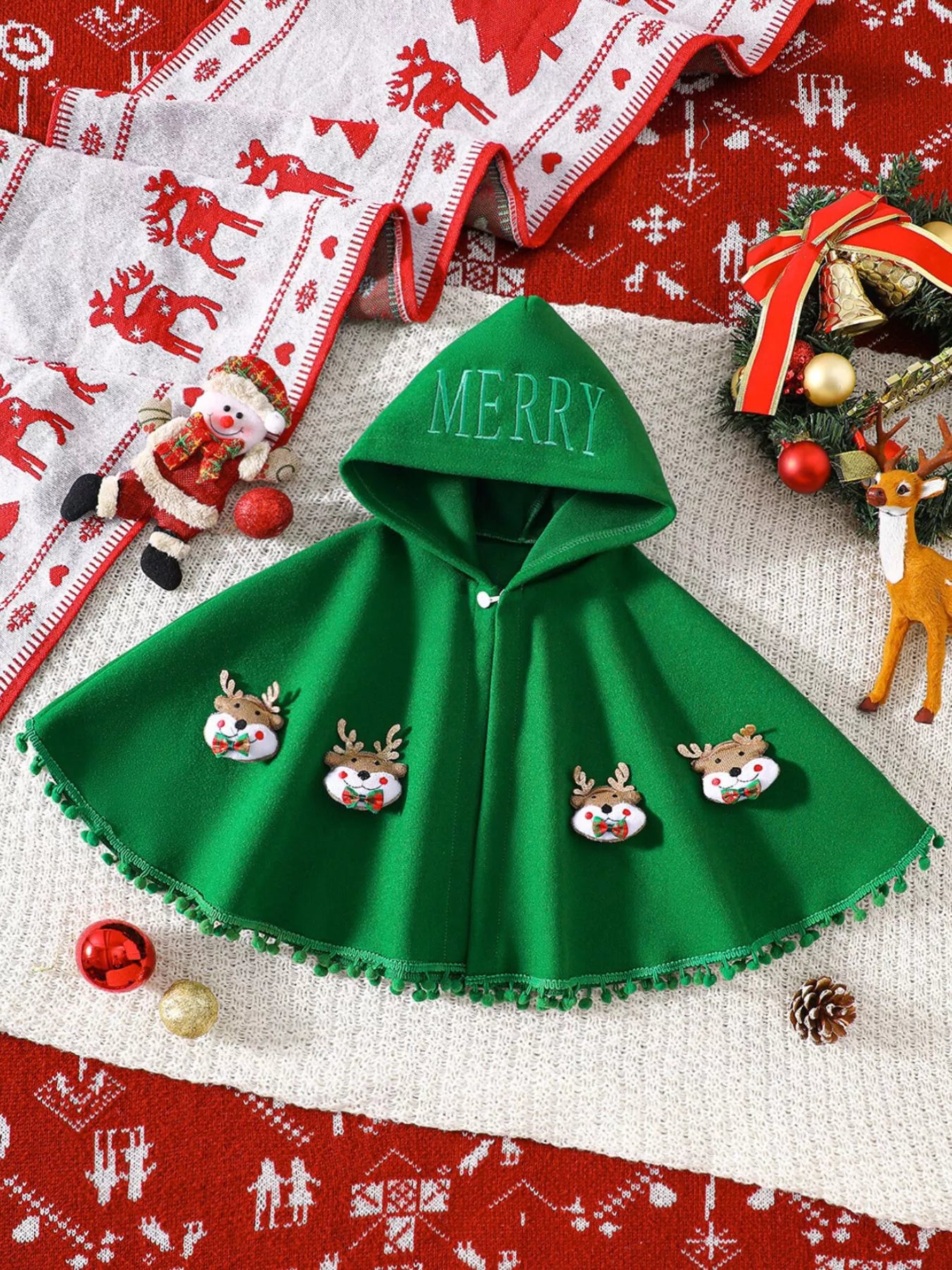 green Christmas hooded cape for kids with “MERRY” embroidery and reindeer appliqués, pom-trim hem, displayed on holiday background.