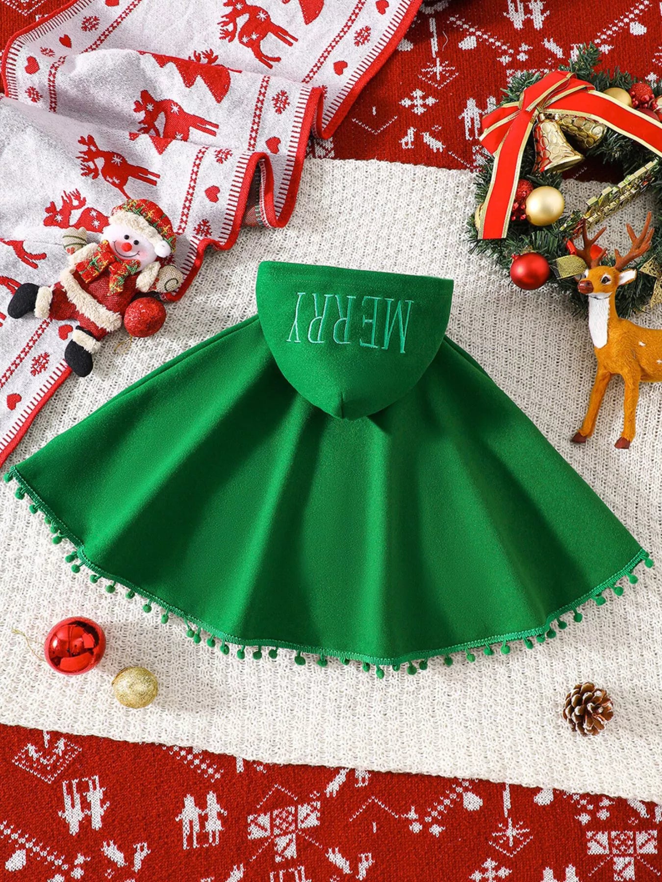 Green Christmas hooded cape for kids with “MERRY” embroidery and reindeer appliqués, pom-trim hem, displayed on holiday background.