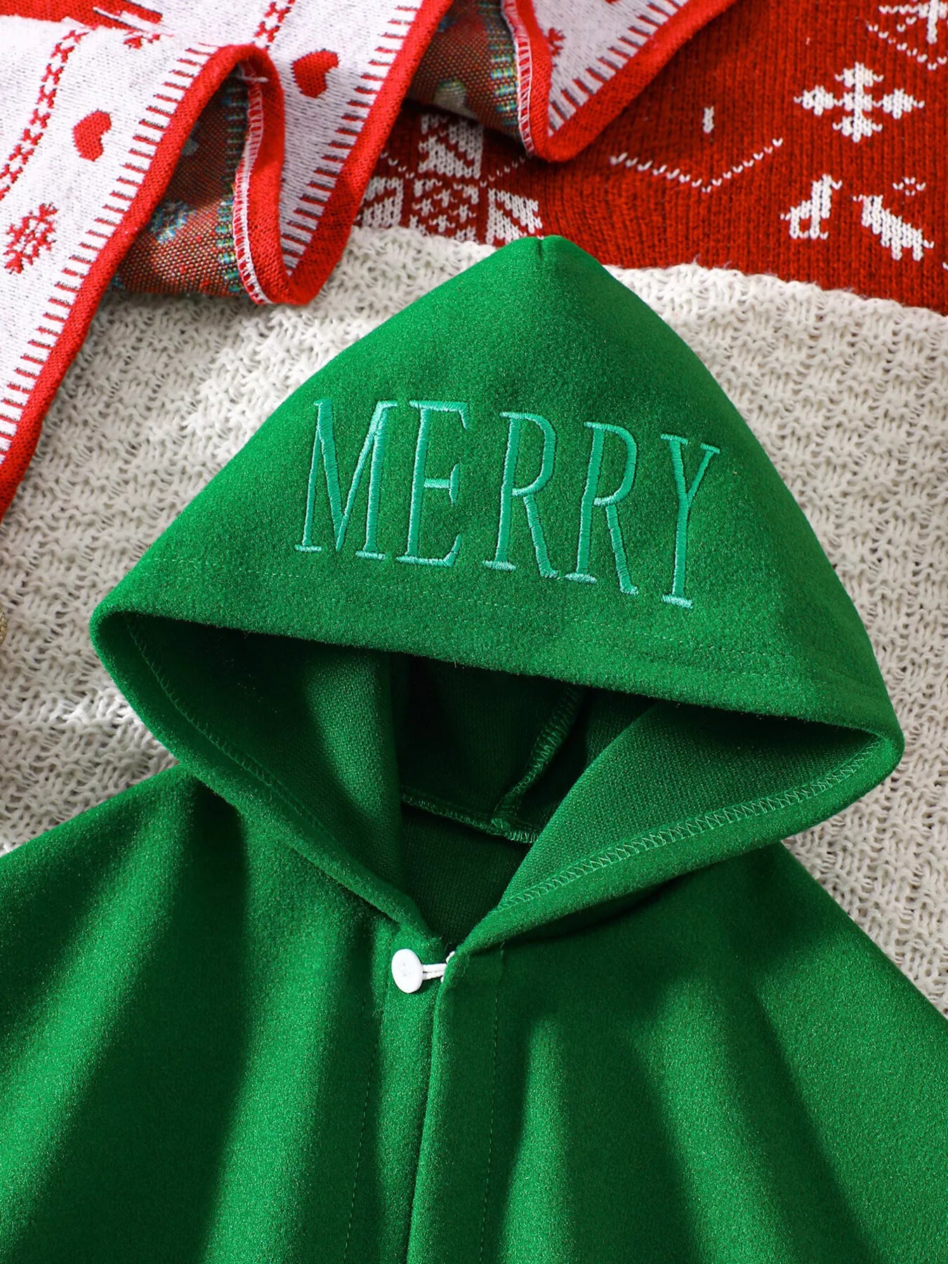 Green Christmas hooded cape for kids with “MERRY” embroidery and reindeer appliqués, pom-trim hem, displayed on holiday background.