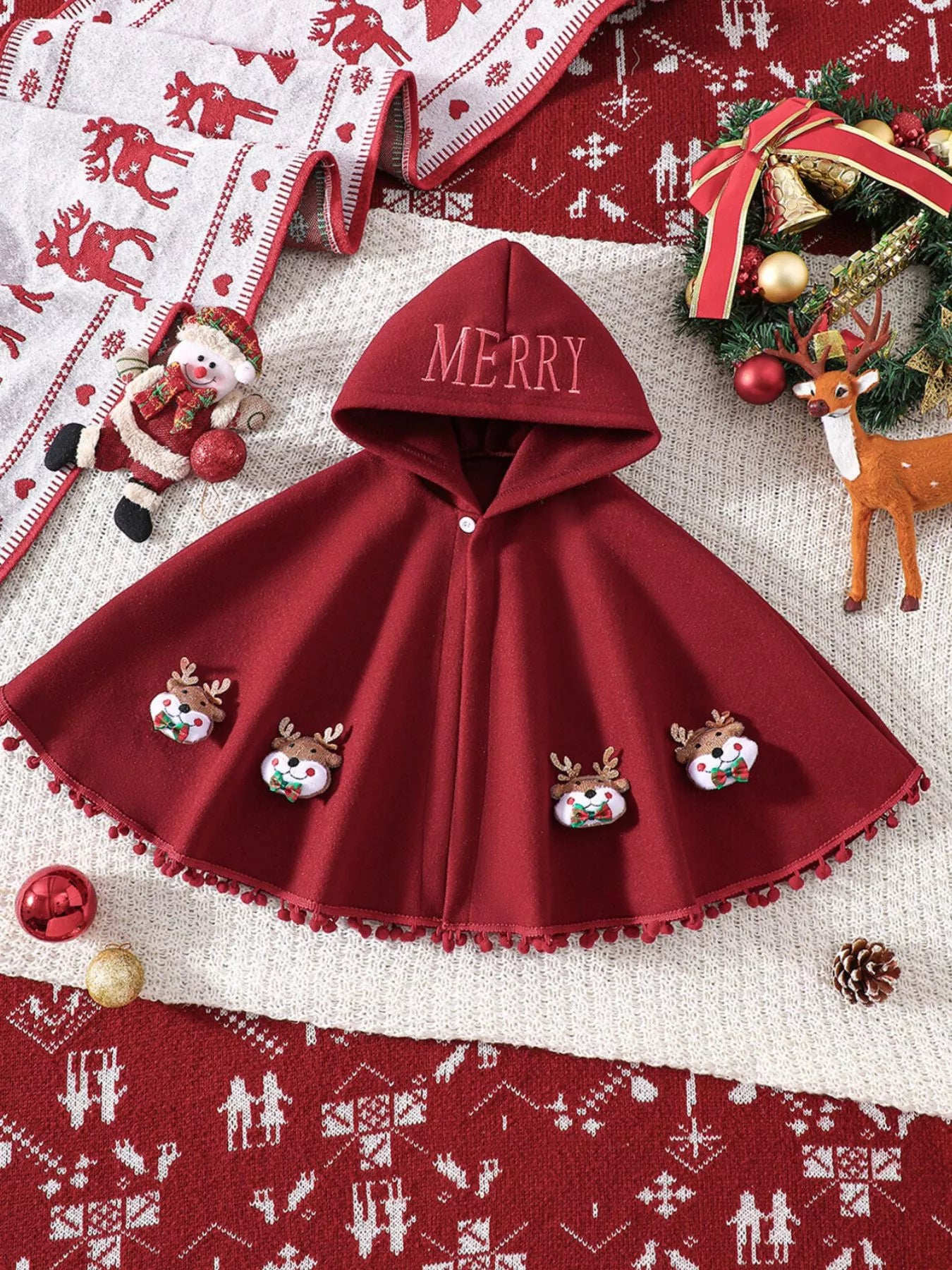 Red Christmas hooded cape for kids with “MERRY” embroidery and reindeer appliqués, pom-trim hem, displayed on holiday background.